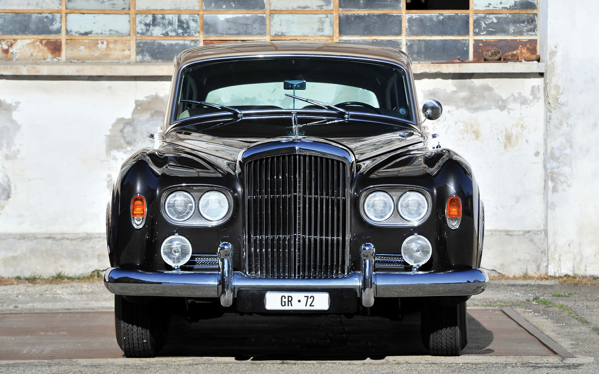 Bentley S3 桌布 壁紙 — 一輛光澤黑色復古賓利，帶有鍍鉻裝飾和圓形頭燈，自豪地矗立在風化的工業牆前。