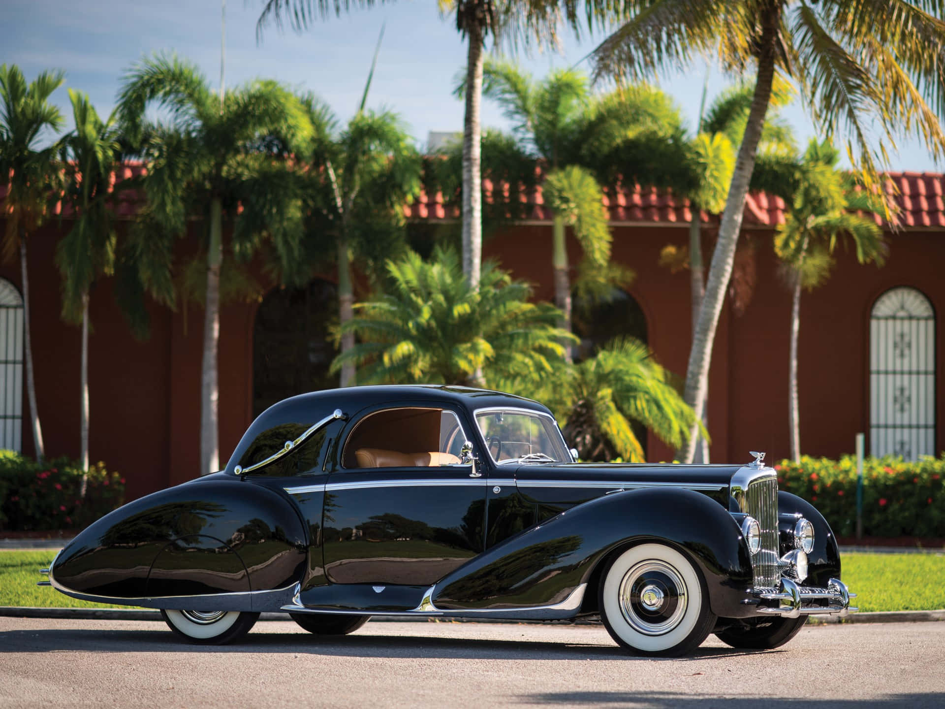 Bentley Mark Vi 桌布 壁紙 — 一輛光澤黑色復古車配白色側壁輪胎，在陽光下閃耀，旁邊有棕櫚樹和一座陶土屋頂建築。