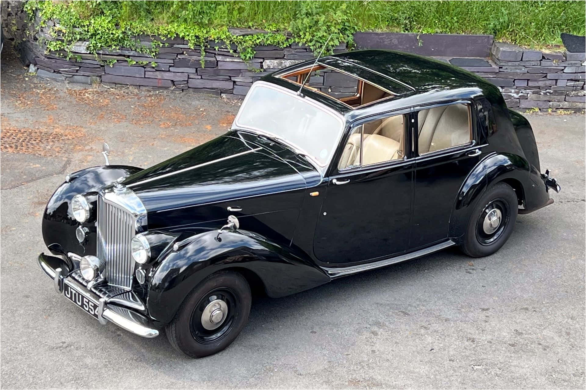 Bentley Mark Vi 桌布 壁紙 — 一輛亮黑色 vintage Rolls-Royce 停放在鋪砌路面上，旁邊是有綠色植物的石牆。