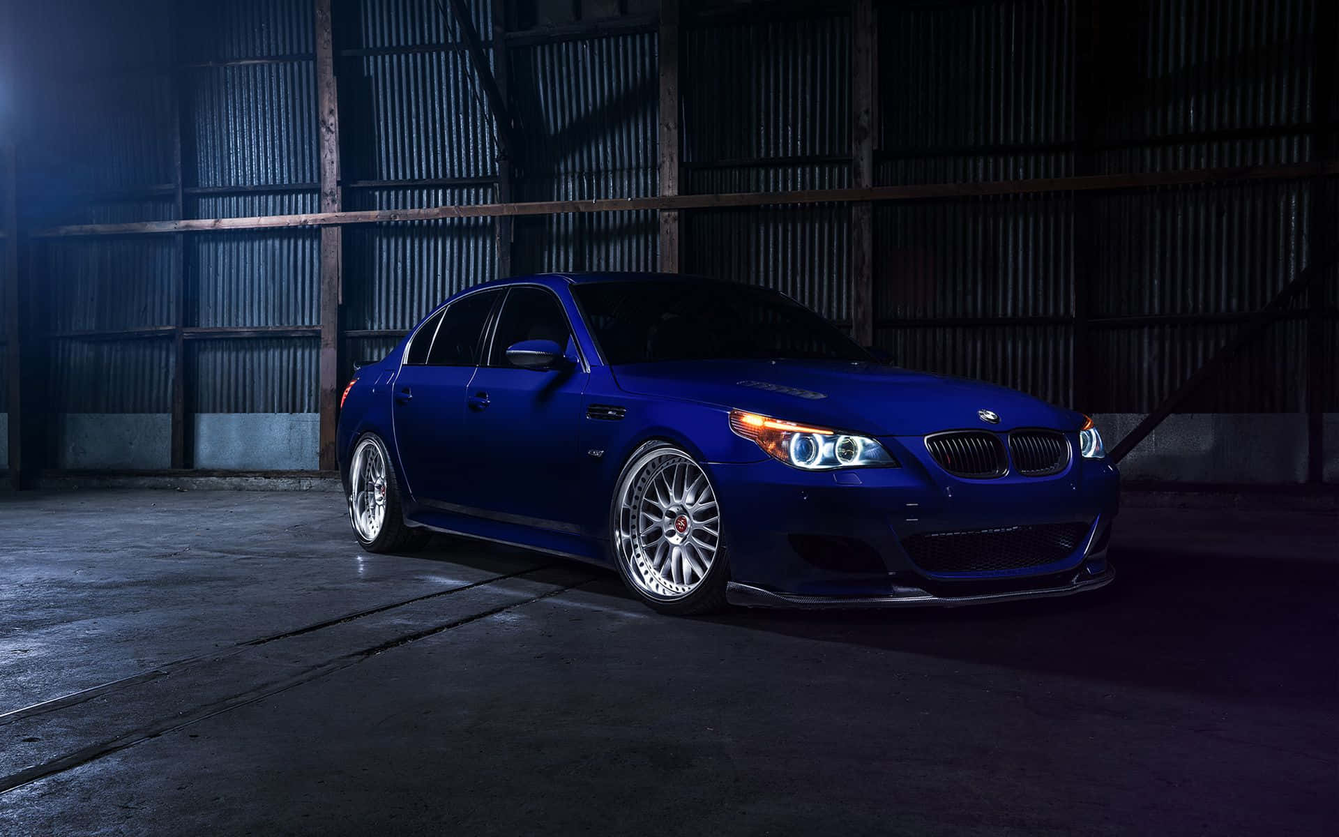 Bmw M5 桌布 壁紙 — 一輛閃亮藍色BMW M5轎車配備定制輪圈，在昏暗的工業倉庫中被戲劇性地照亮。