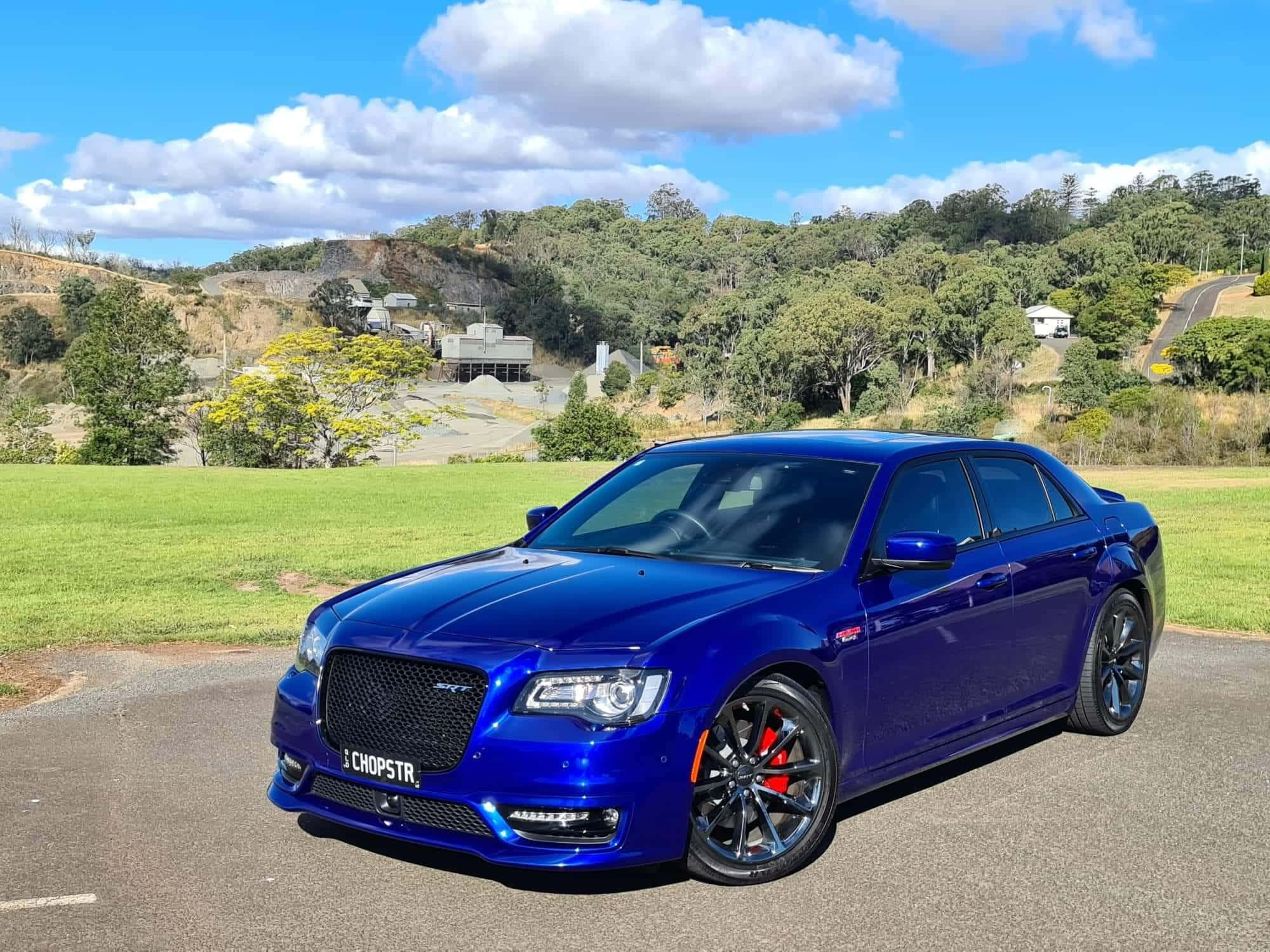 Chrysler 300 桌布 壁紙 — 一輛閃亮的藍色肌肉車停在柏油路上，背景有綠色山丘和散落的房屋，上方是晴朗的藍天和蓬鬆的白雲。