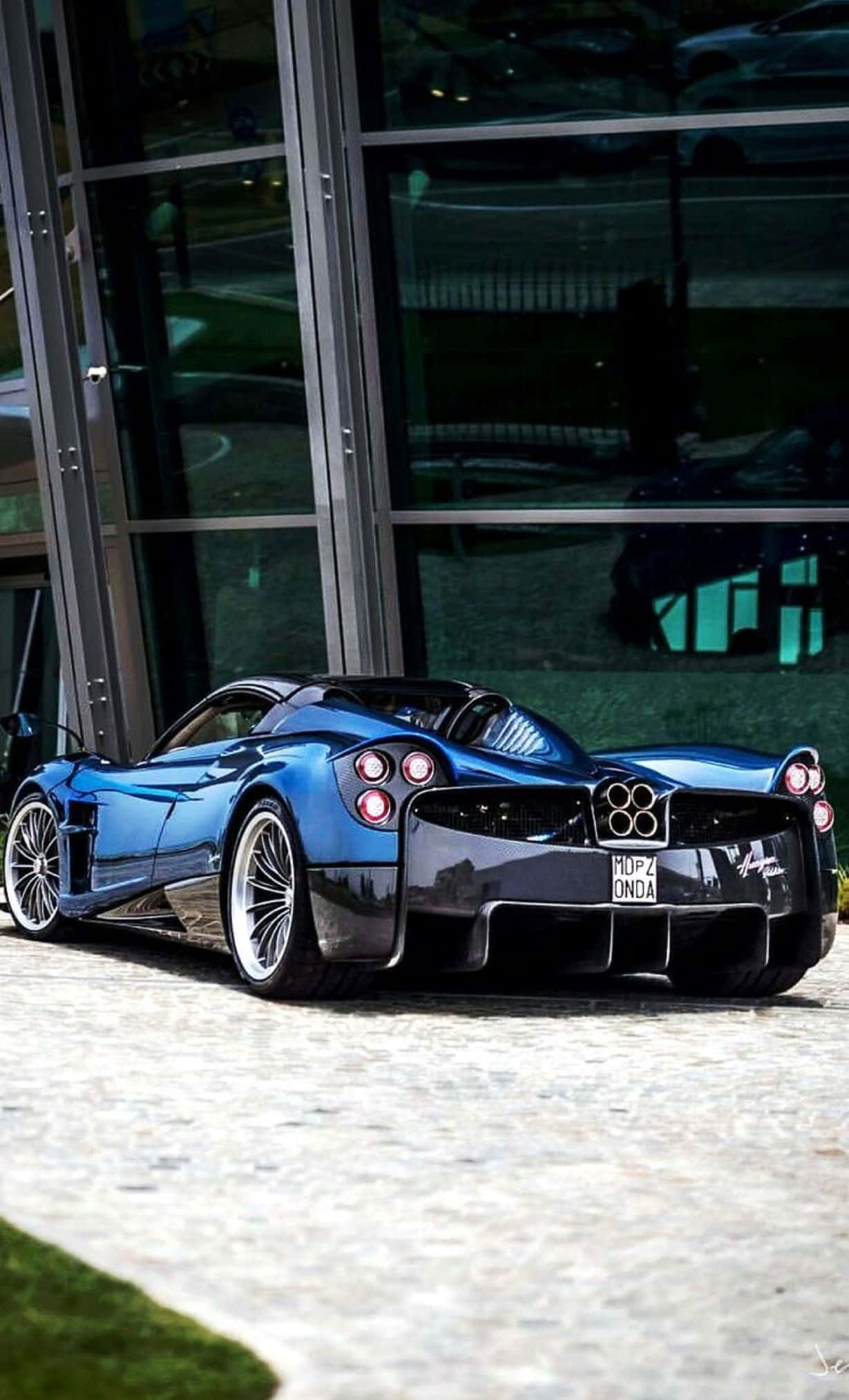 Pagani Huayra Roadster Bc 桌布 壁紙 — 一輛閃亮藍色Pagani Huayra跑車停放在現代玻璃建築旁的石鋪路面上。