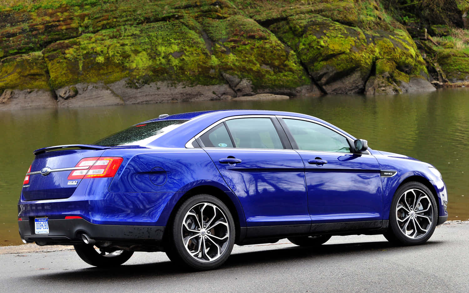 Ford Taurus 桌布 壁紙 — 一輛光澤的藍色轎車停泊在長滿苔蘚的河岸旁，水面反射，地形多石。