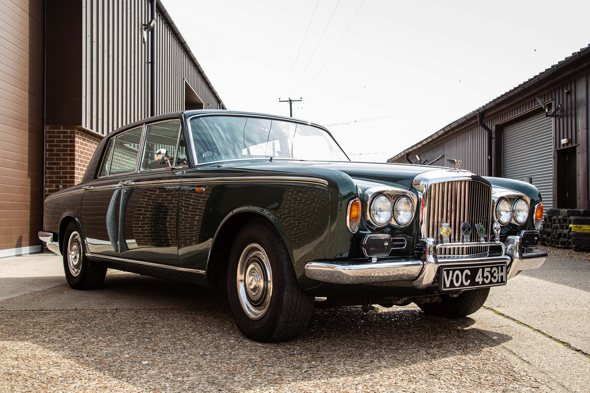 Bentley T1 桌布 壁紙 — 一輛光澤深綠色復古賓利轎車停放在工業建築之間的礫石上，在明亮的日光下。
