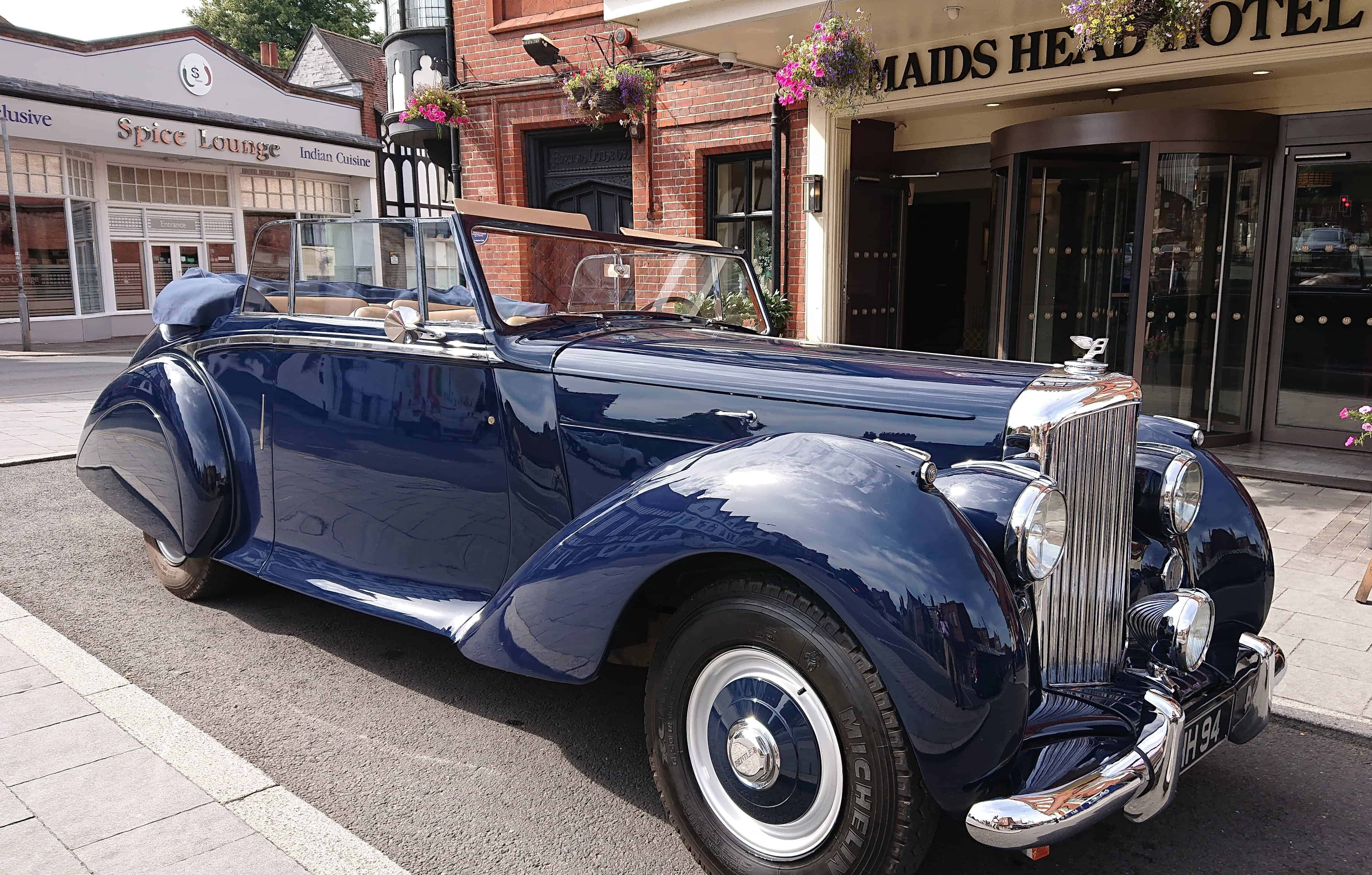 Bentley Mark Vi 桌布 壁紙 — 一輛光澤深藍色復古敞篷車停在鵝卵石街道上，前面是一間有掛花籃的磚砌酒店。