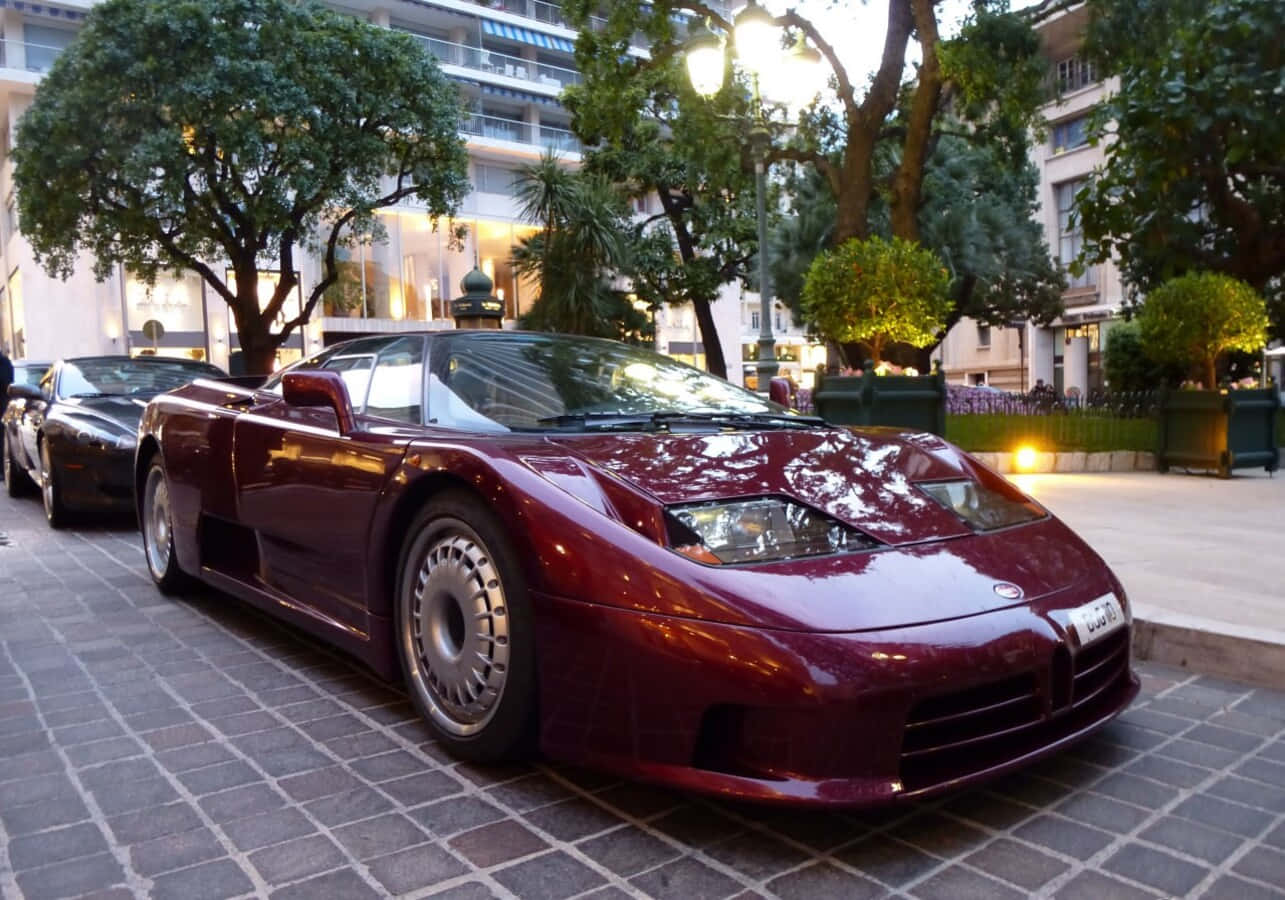 Bugatti Eb110 桌布 壁紙 — 一輛光澤的栗色布加迪EB110超級跑車在街燈下閃閃發光，停在一個被樹木和建築物包圍的鵝卵石城市廣場上。