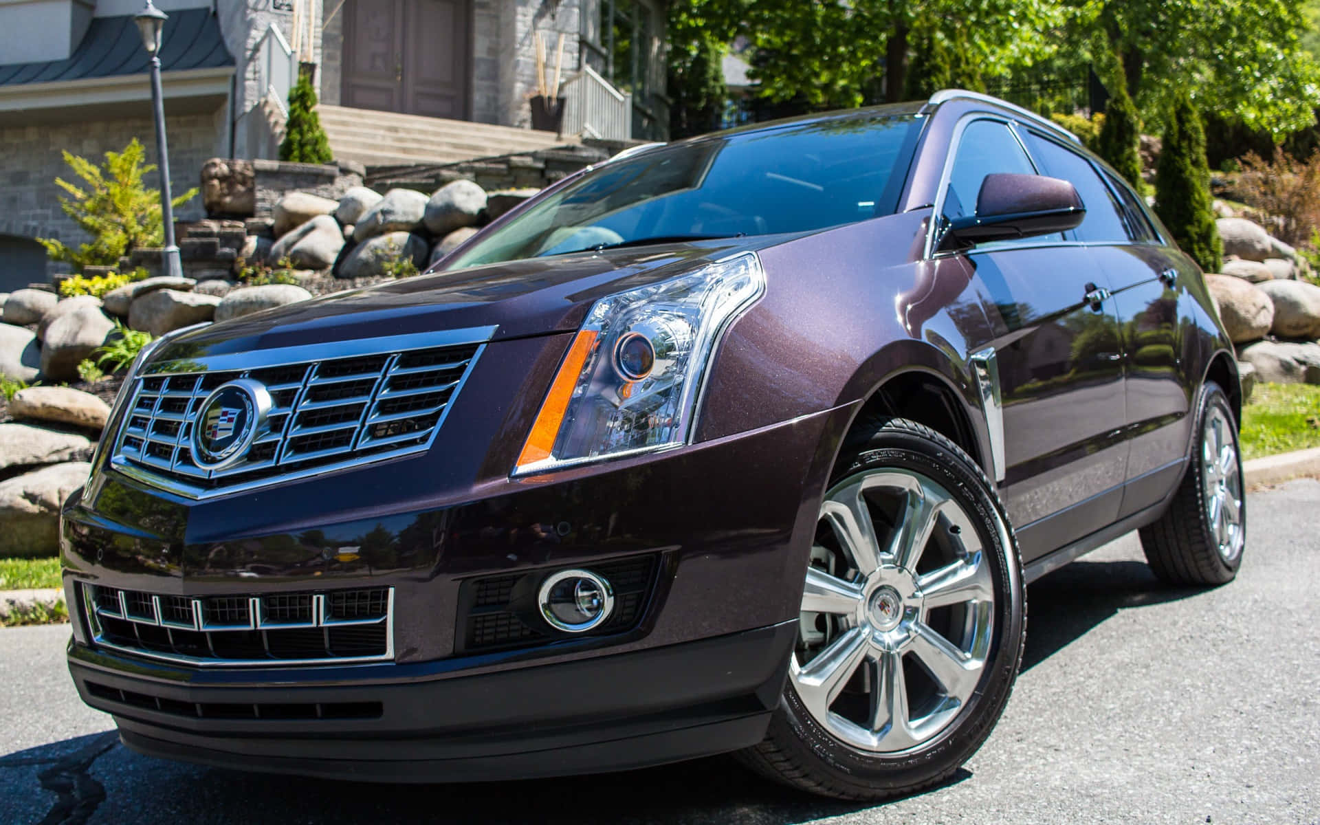 Cadillac Srx 桌布 壁紙 — 一輛光澤的栗色Cadillac SRX SUV停放在陽光明媚的住宅街道上，周圍有石頭景觀和綠樹。