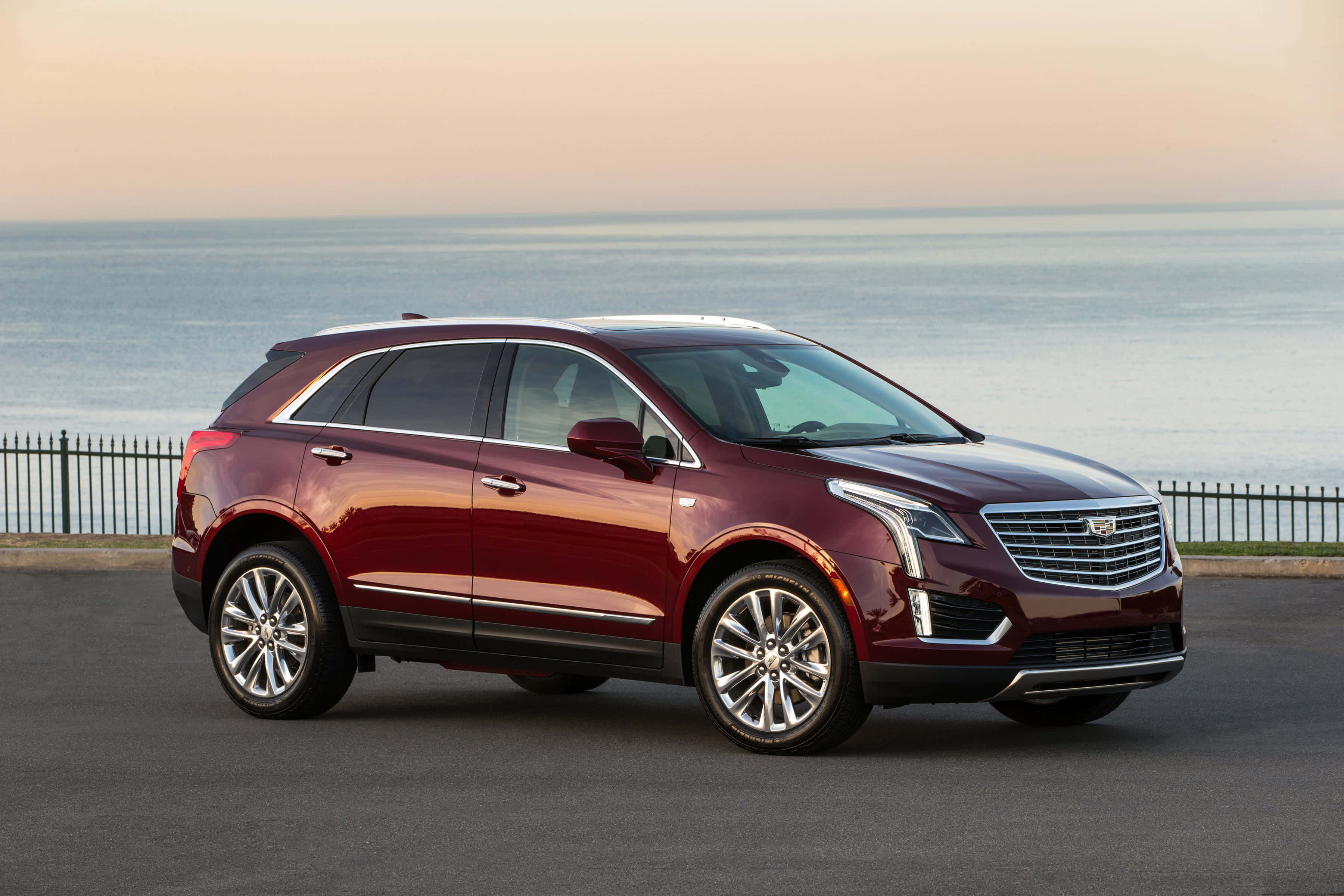 Cadillac Xt5 桌布 壁紙 — 一輛光澤的栗色凱迪拉克SUV在日落時分停泊在海邊，背後是平靜的水面和柔和的天色。
