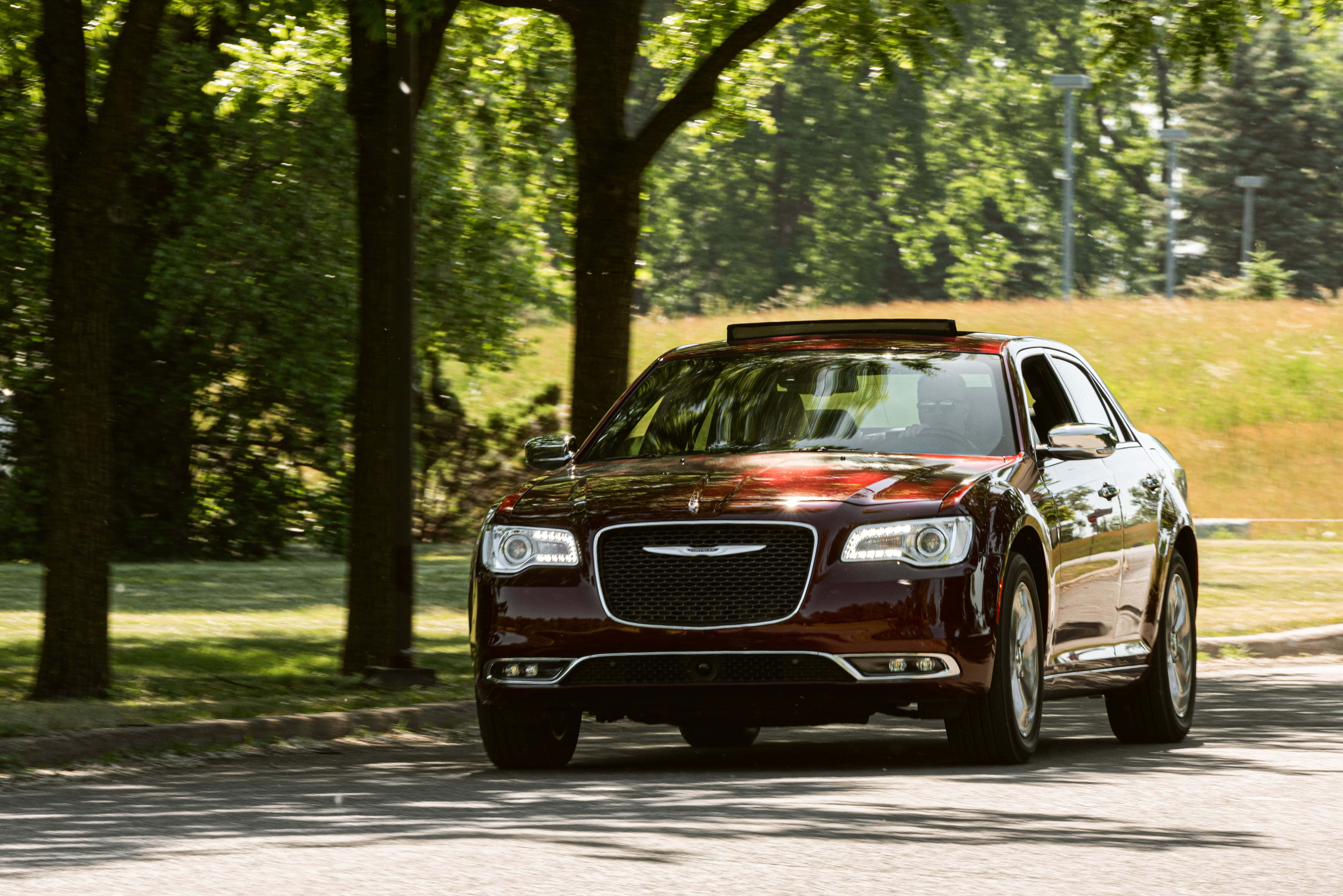 Chrysler 300 桌布 壁紙 — 一輛光澤的栗色豪華轎車在明亮的夏日陽光下行駛在綠樹成蔭的公園道路上。