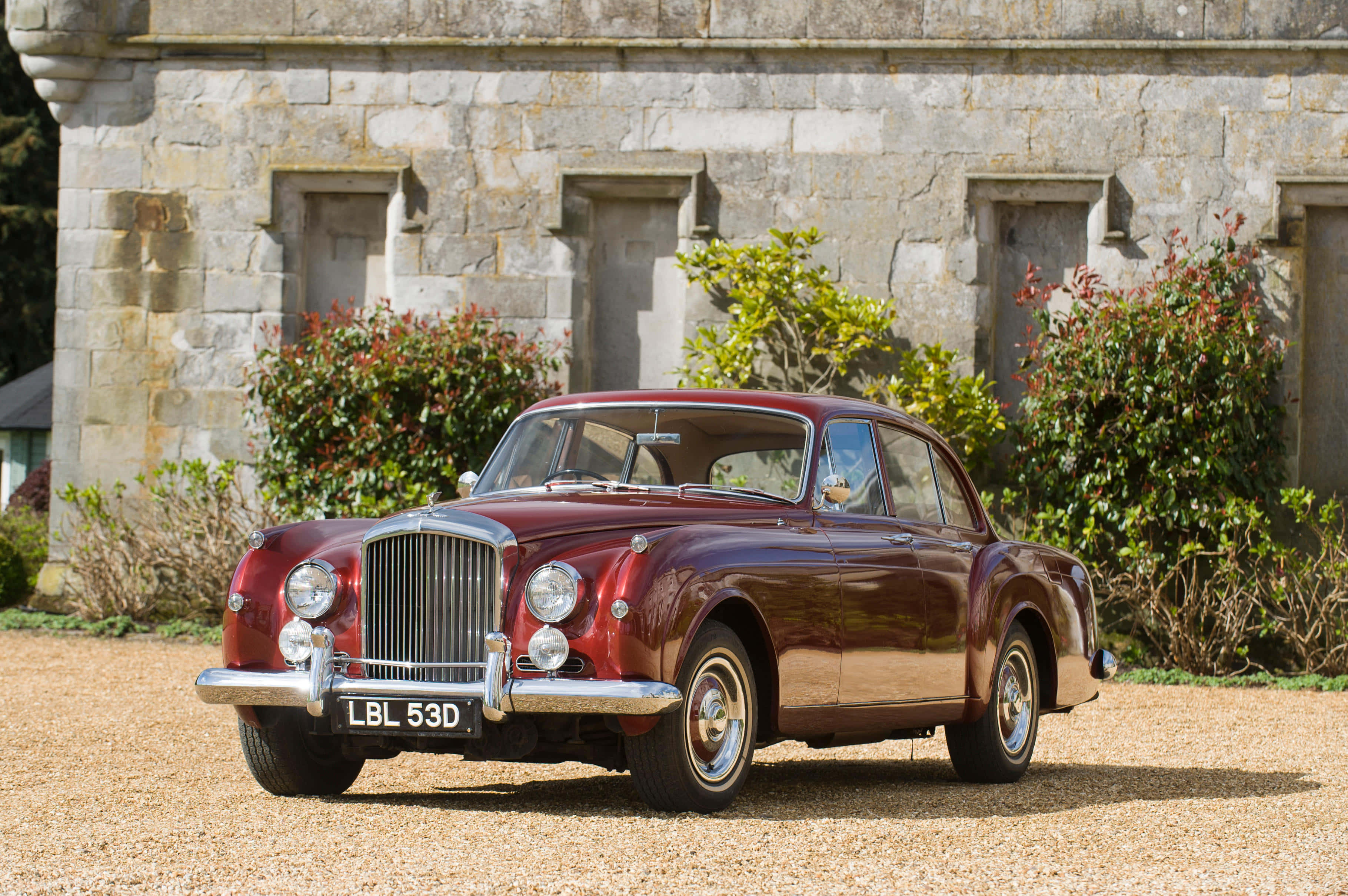 Bentley S2 桌布 壁紙 — 一輛亮面栗色復古賓利房車停泊在碎石路上，前方是有綠色植物的石建築物。