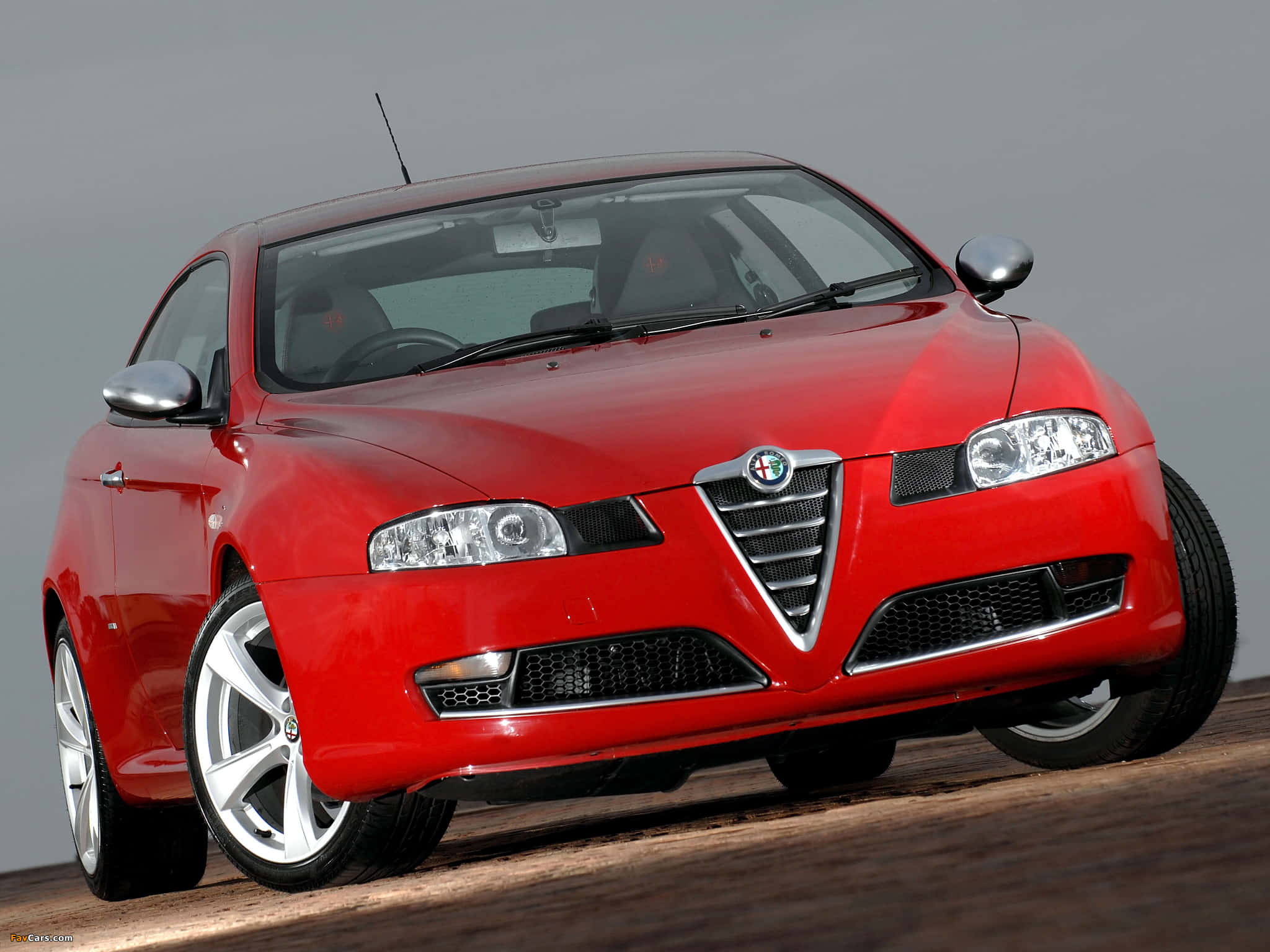 Alfa Romeo Gt 桌布 壁紙 — 一輛亮紅色 Alfa Romeo 運動型轎跑車在木質表面上以正面角度擺放，背景為中性灰色。