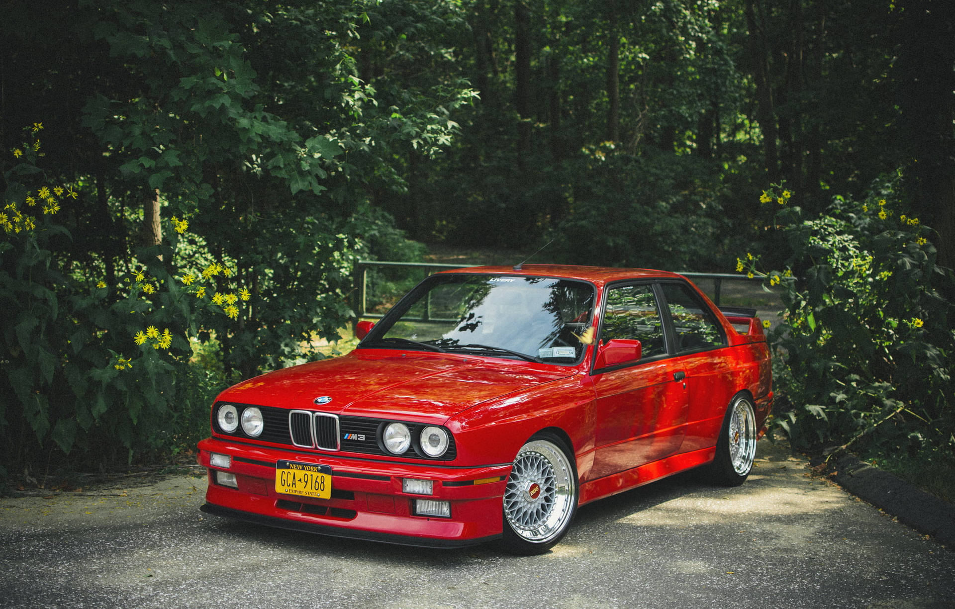 紅色BMW E30 M3停泊在夏日森林的碎石路上，伴有黃色花朵。 — 跑車 壁紙