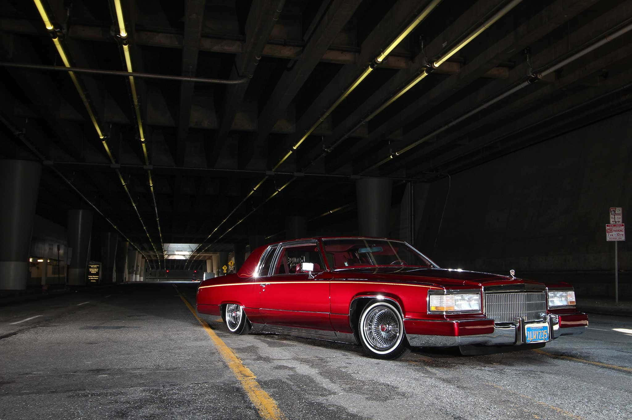 Cadillac Deville 桌布 壁紙 — 一輛光澤的紅色經典凱迪拉克轎車配備定制輪圈，停放在昏暗的城市高架橋下，帶有黃色燈光。