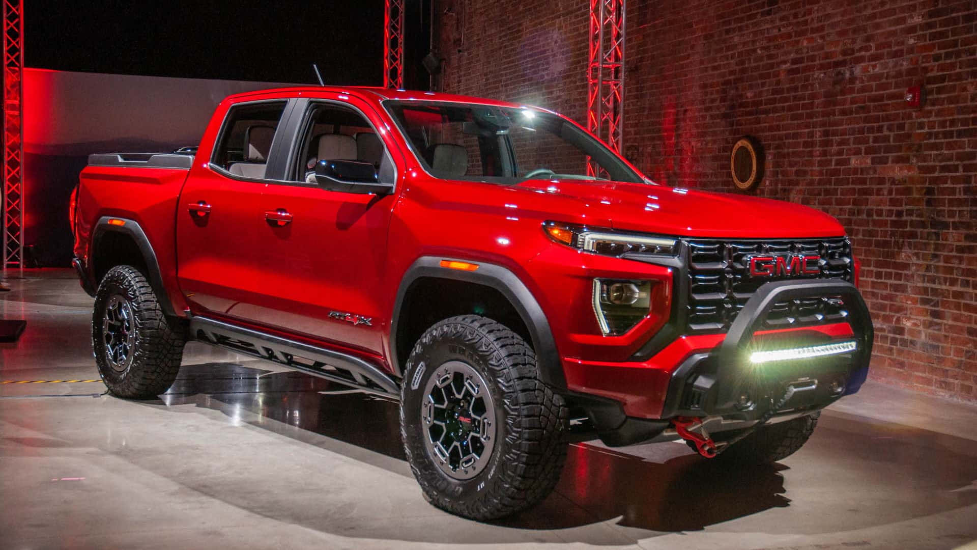 Gmc Canyon 桌布 壁紙 — 一輛帶有黑色飾邊和堅固輪胎的光澤紅色GMC Sierra AT4皮卡車，在戲劇性的紅色燈光下，以磚牆為背景展示。