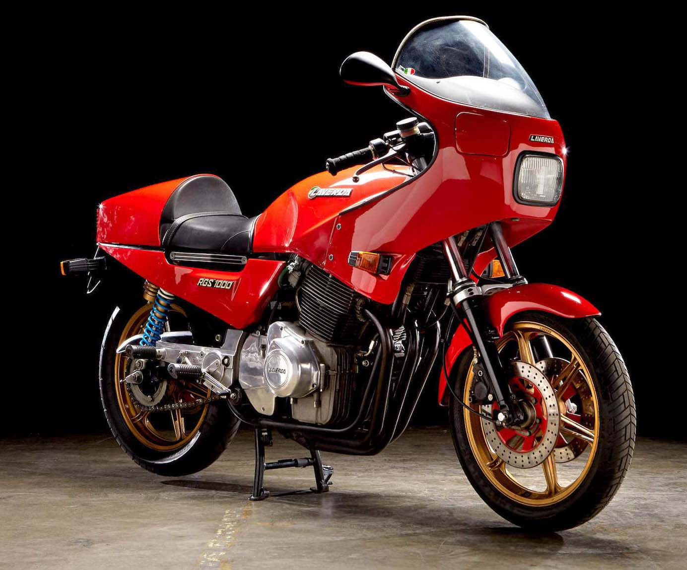  壁紙 — 一輛光澤紅色 Kawasaki ZGS 1000 摩托車，配金色輪圈和藍色懸吊，以側腳架停放在純黑背景前。