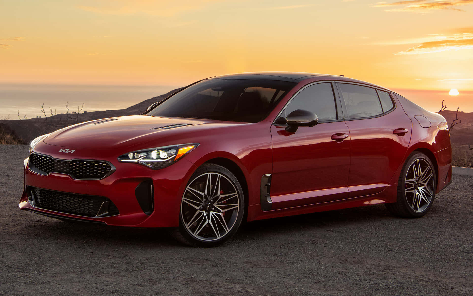 Kia Stinger 桌布 壁紙 — 一輛亮紅色的Kia Stinger運動轎車停在日落時分的碎石觀景點，背景是橙色天空和遠方山丘。