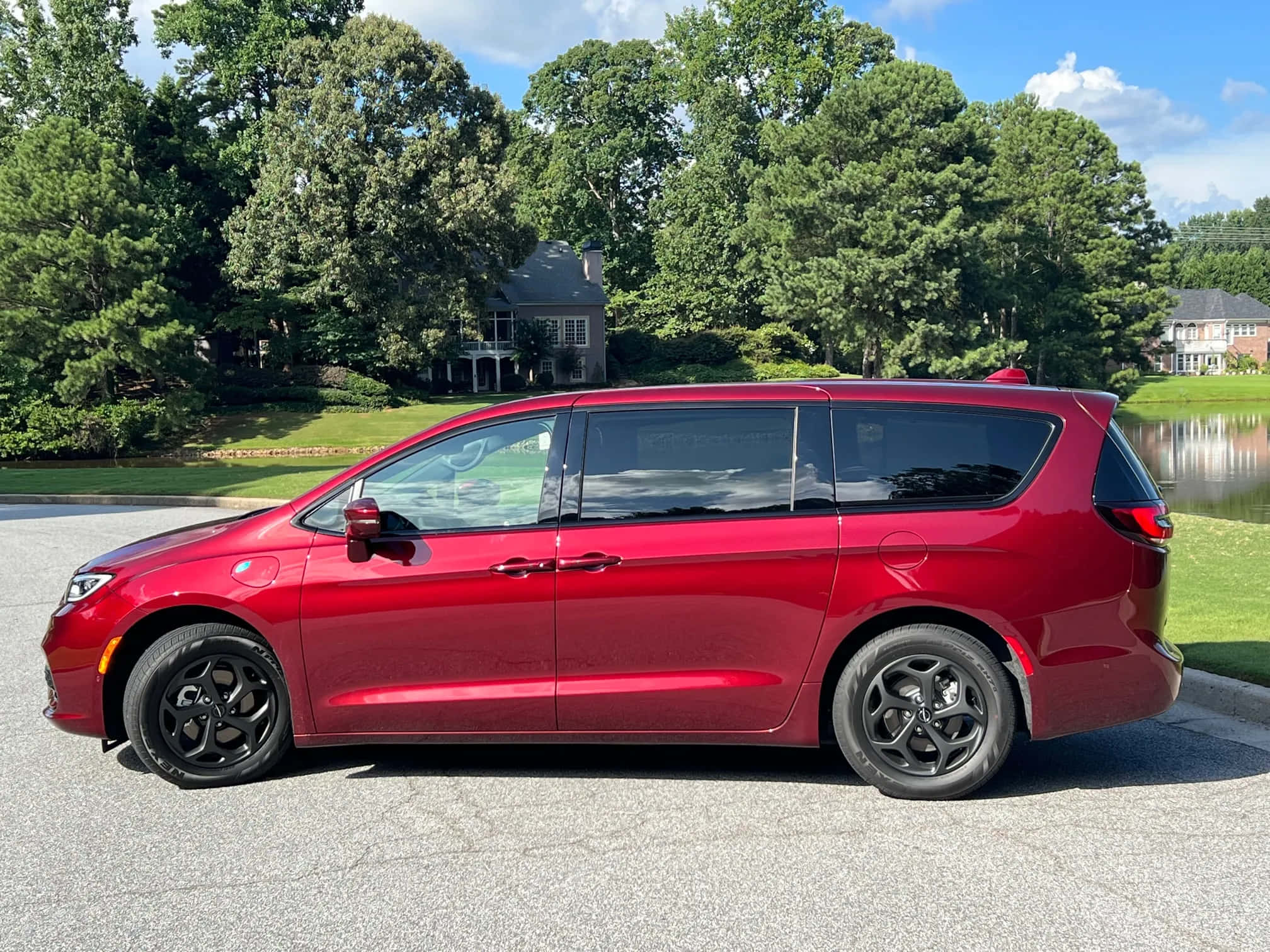 Chrysler Pacifica 桌布 壁紙 — 一輛亮紅色小型貨車配黑色輪圈，停放在鋪砌車道上，旁邊是寧靜的湖泊和茂密的綠樹，天空湛藍。