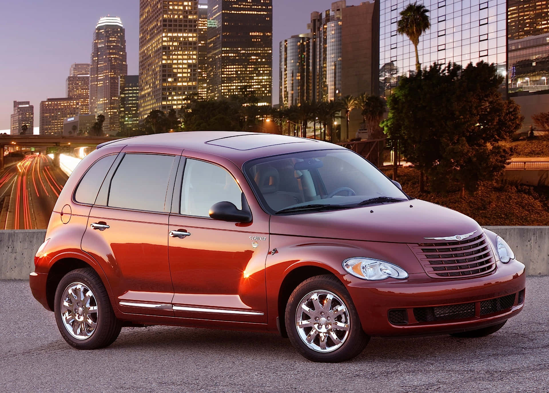 Chrysler Pt Cruiser 桌布 壁紙 — 一輛亮紅色的PT Cruiser停在城市街道上，有模糊的交通痕跡和燈光閃爍的摩天大樓在黃昏時分。