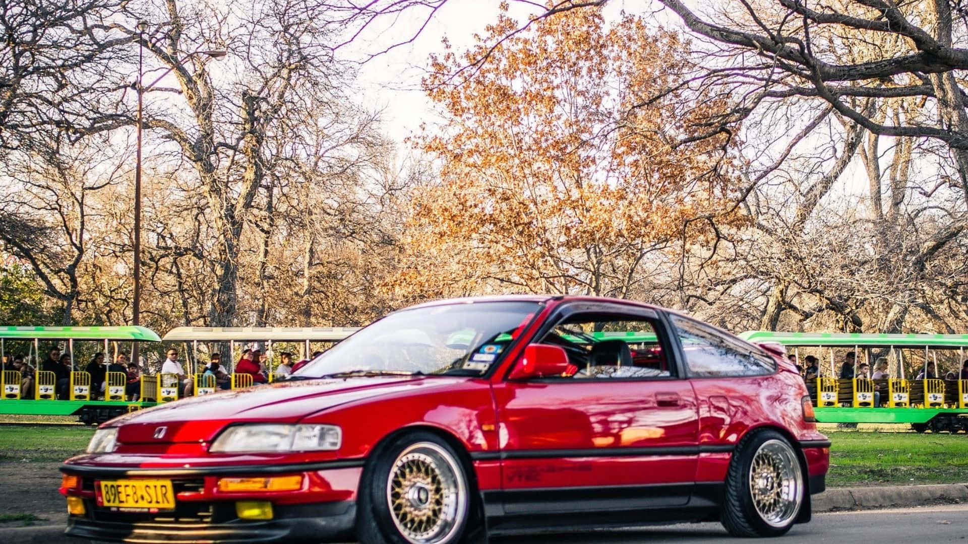Honda Civic Del Sol 桌布 壁紙 — 一輛光亮的紅色跑車停放在公園裡，背景有光禿的樹木和綠色電車。