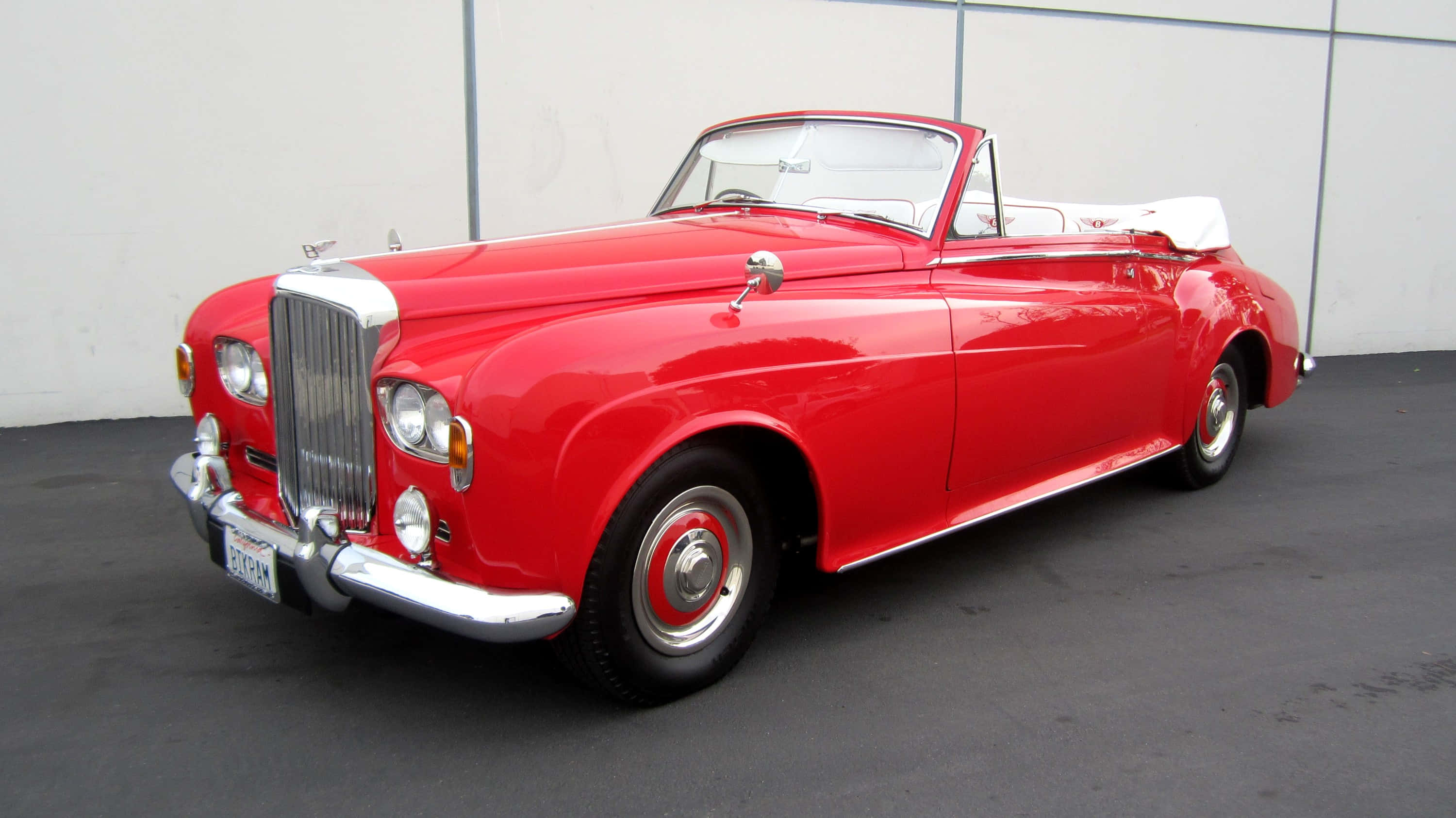 Bentley S3 桌布 壁紙 — 一輛光澤的紅色復古敞篷跑車，配備白色內裝和鍍鉻裝飾，停放在純白色牆壁前。