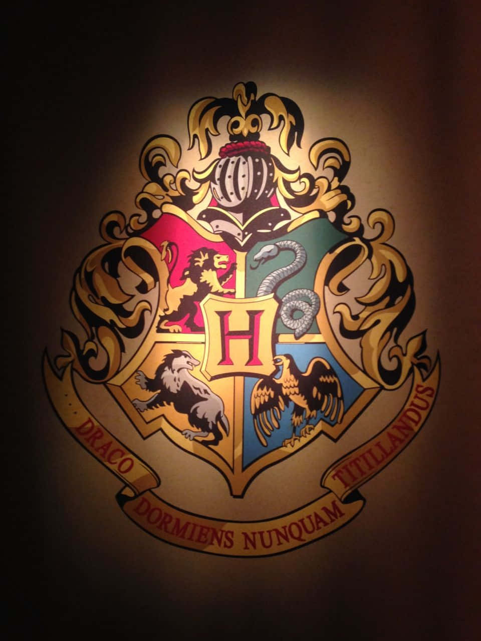 Hogwarts Crest 桌布 壁紙 — 一個發光的霍格華茲徽章，在聚光燈下映照在深色牆壁上，包含四個學院的標誌和格言。