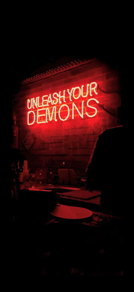  壁紙 — 一個發光的紅色霓虹燈牌，上面寫著 unleash your demons，在燈光昏暗的酒吧中，在磚牆上投射出詭異的光芒。
