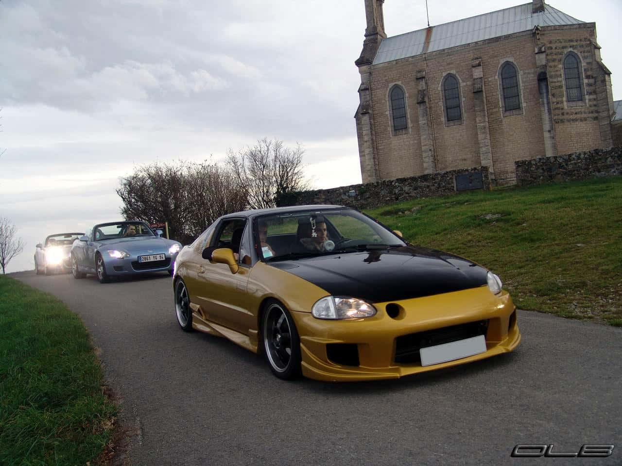 Honda Civic Del Sol 桌布 壁紙 — 一輛金色和黑色的改裝跑車在陰天下行駛在一條蜿蜒的道路上，經過一座石頭教堂。