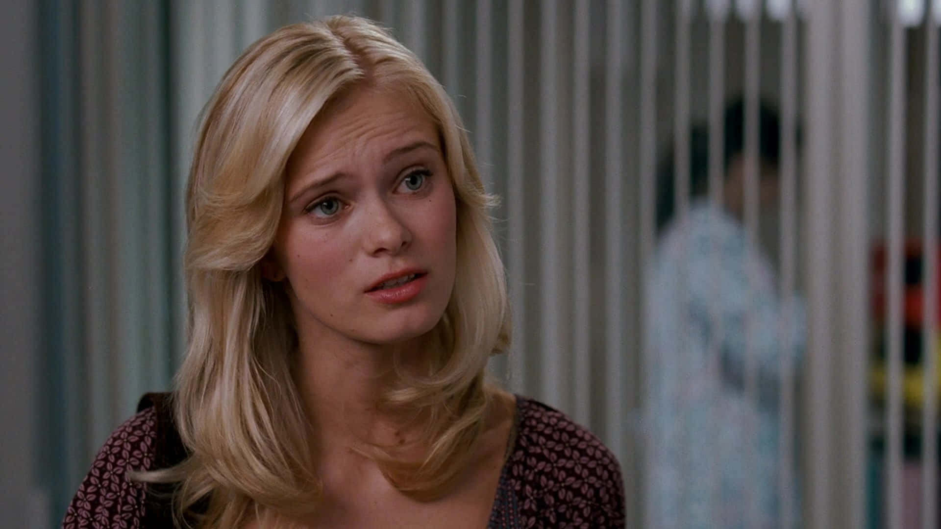 Sara Paxton 桌布 壁紙 — 一位金髮女子帶著憂慮的表情望向鏡頭外，身處一個光線柔和、有垂直百葉窗的室內場景。
