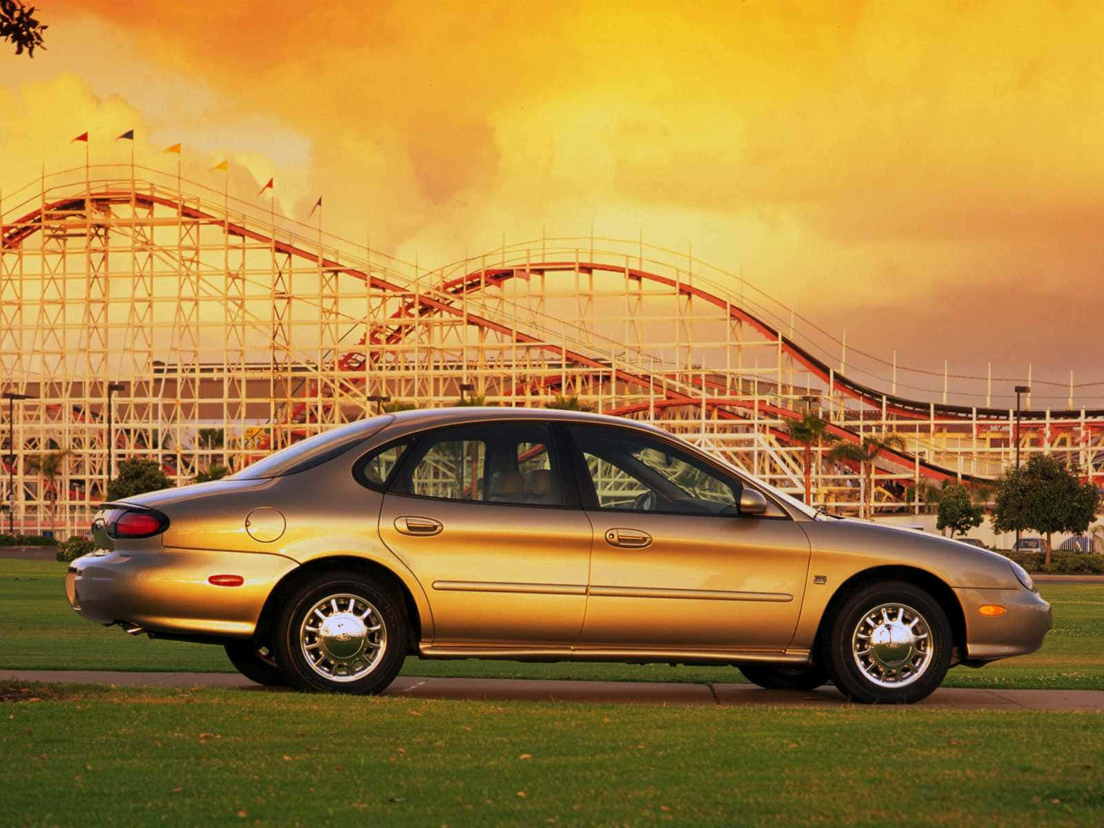 Ford Taurus 桌布 壁紙 — 一輛金色轎車停在草地上，高聳的雲霄飛車在火紅的日落天空下閃耀。