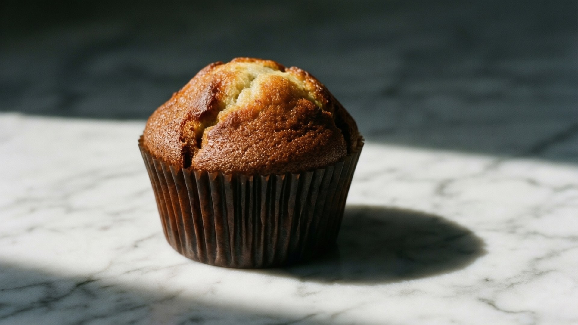 Minimalist Muffin 壁紙 — 一個金黃色的馬芬蛋糕放在深色紙托上，置於白色大理石表面，沐浴在明亮的陽光下，投射出清晰的陰影。