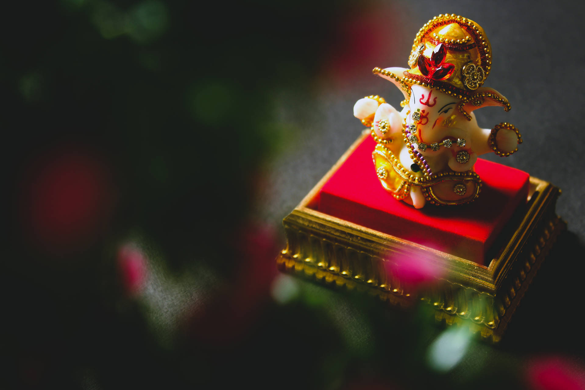 金色象神雕像在紅色軟墊底座上，帶模糊花卉背景。 — 神 壁紙