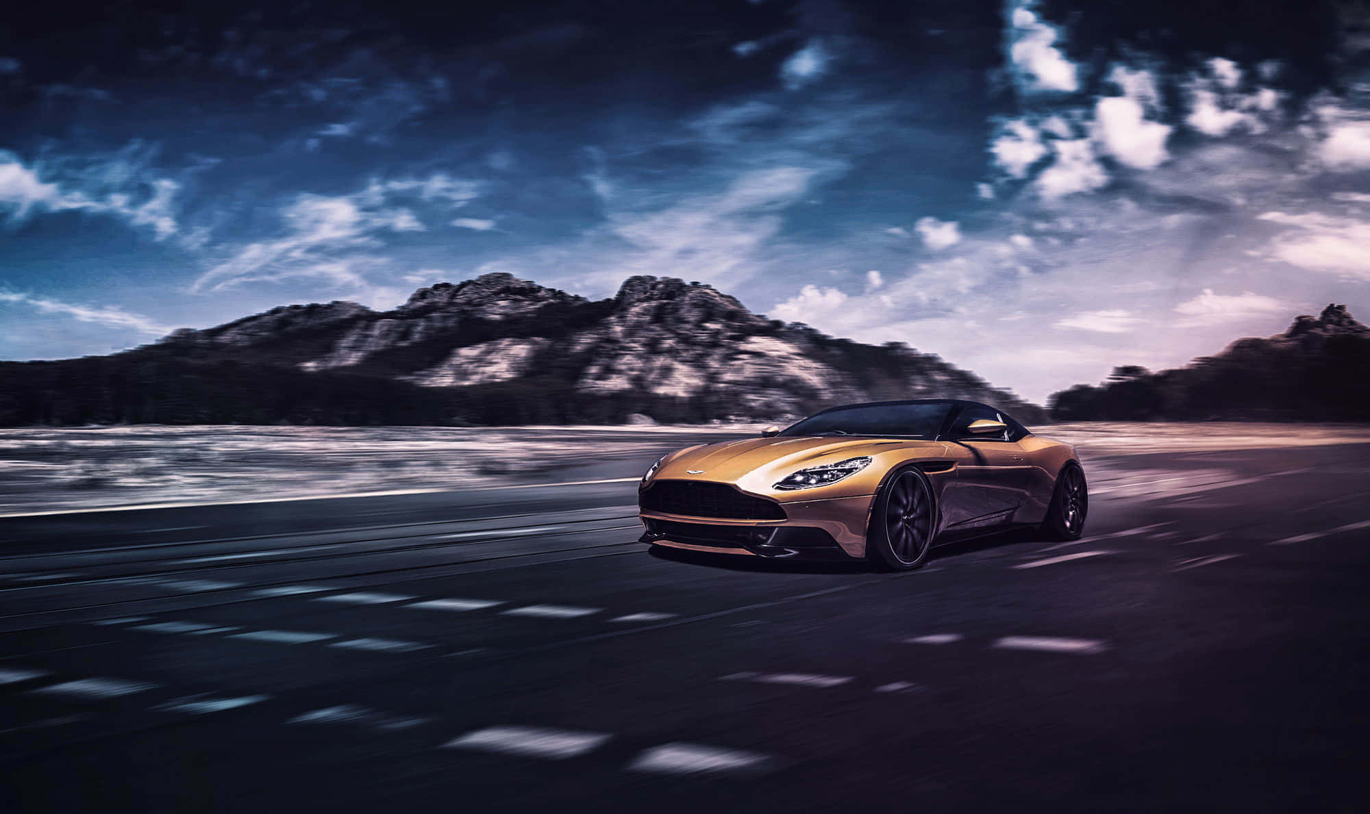 Aston Martin Db11 桌布 壁紙 — 一輛金色跑車在昏暗的道路上疾馳，帶有模糊的動感，背景是戲劇性的暮色天空和遠山。