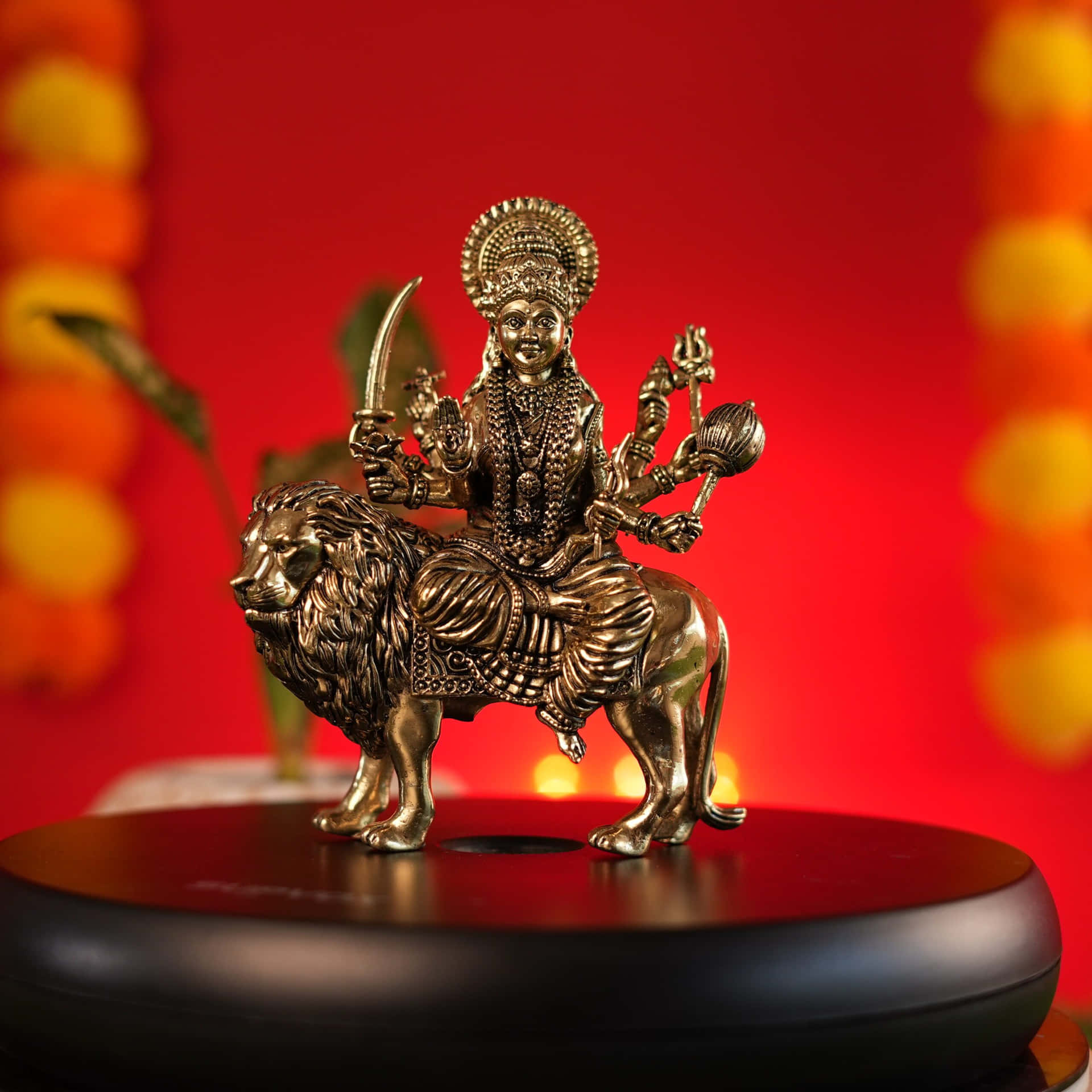  壁紙 — 金色的杜爾迦女神騎著獅子的雕像，背景是鮮紅色，還有模糊的萬壽菊花環。