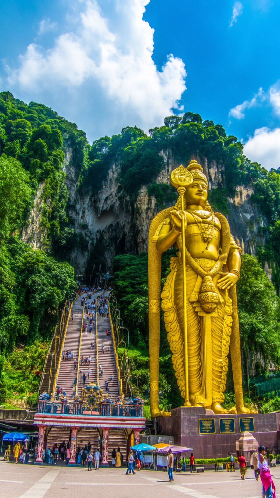 Batu Caves Golden Deity 壁紙 — 一座高聳的黃金穆魯甘神像矗立在鬱鬱蔥蔥的綠色懸崖前，長長的樓梯通向洞穴入口，上方是明亮的藍天。