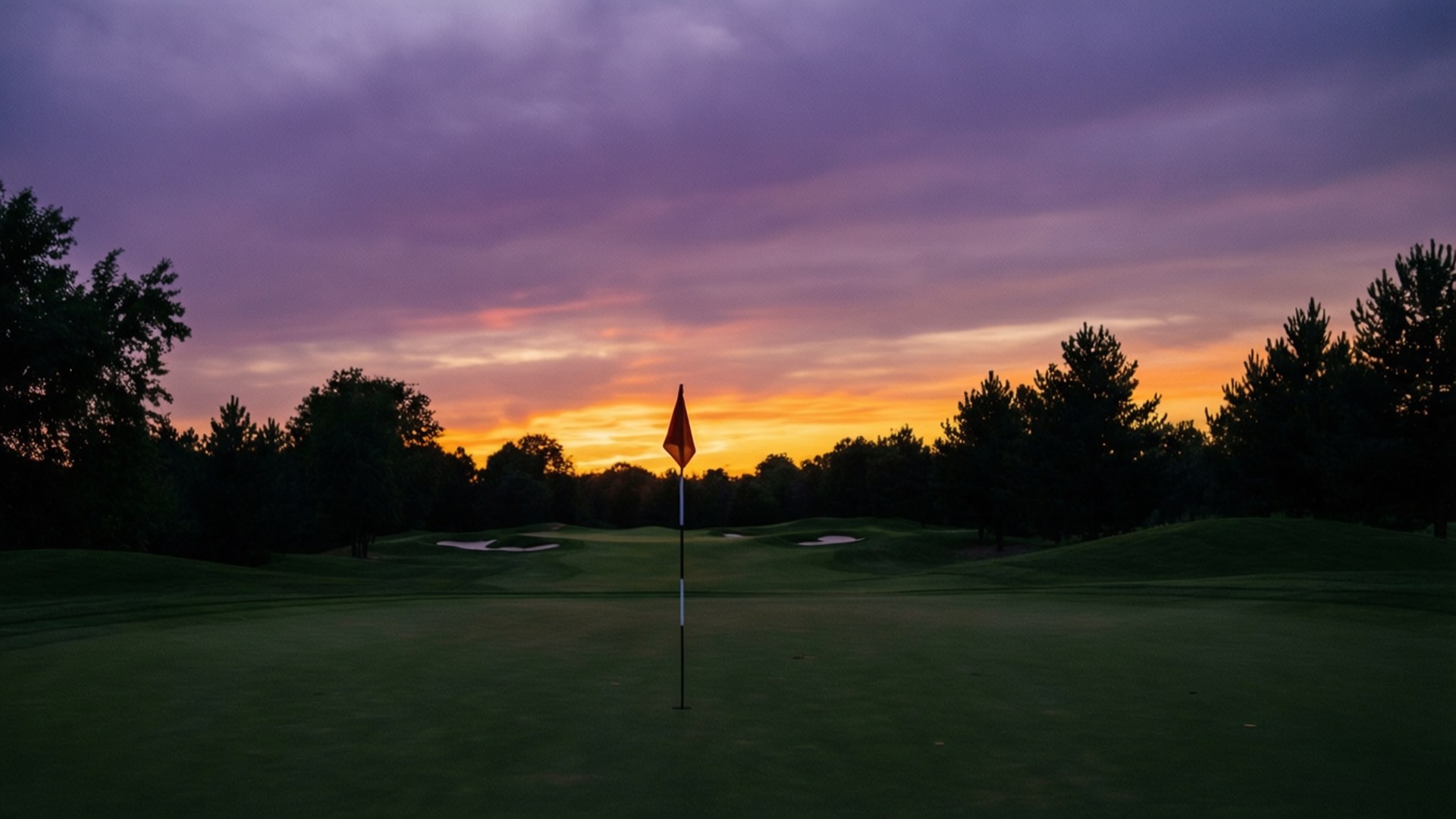 Dusk on the Course 壁紙 — 一支高爾夫球旗獨自立在果嶺上，鮮豔的紫色和橙色夕陽在剪影樹木後方描繪天空。