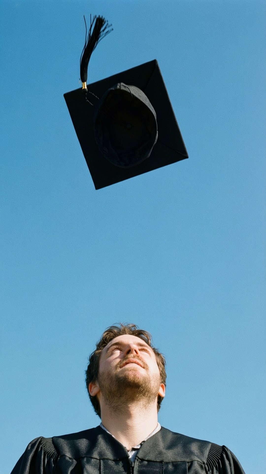 Graduation Cap Toss 壁紙 — 一位身穿黑色畢業袍的畢業生，帶著勝利的表情，將學士帽高高拋向清澈的藍天。