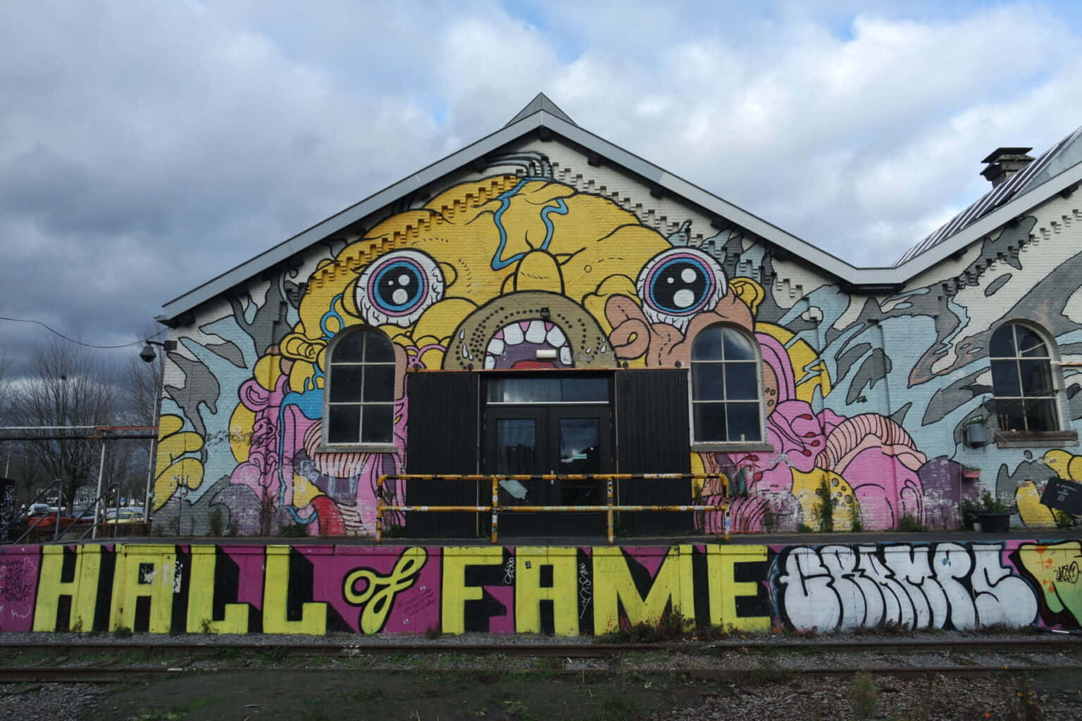 Tilburg 桌布 壁紙 — 一個充滿活力的塗鴉倉庫，上面有巨大的卡通臉壁畫和「HALL OF FAME」文字，在陰天天空下。