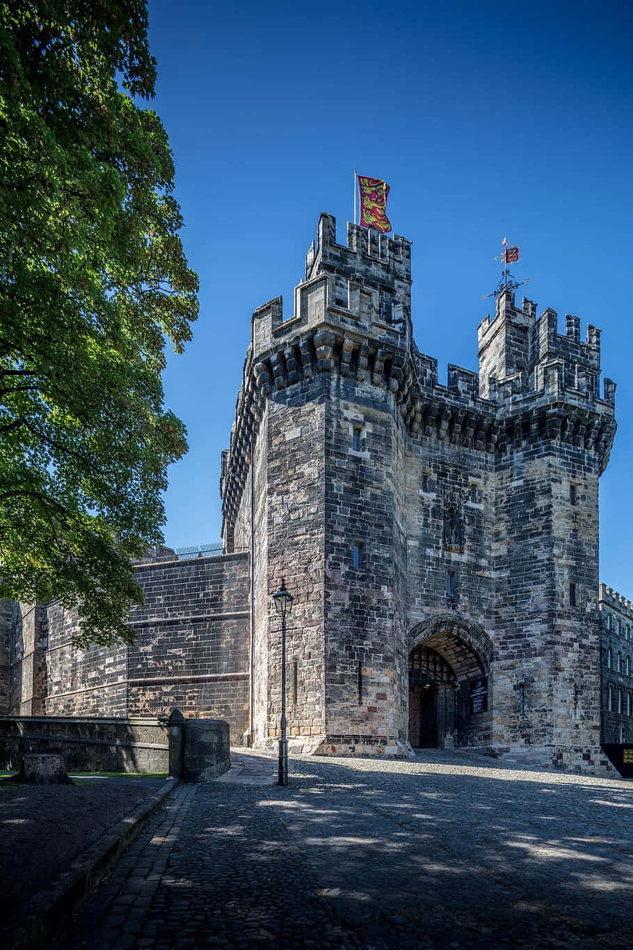 Lancaster United Kingdom 桌布 壁紙 — 一座宏偉的中世紀石造城堡大門，帶有城垛塔樓和旗幟，在晴朗的藍天下，旁邊是茂密的綠樹。