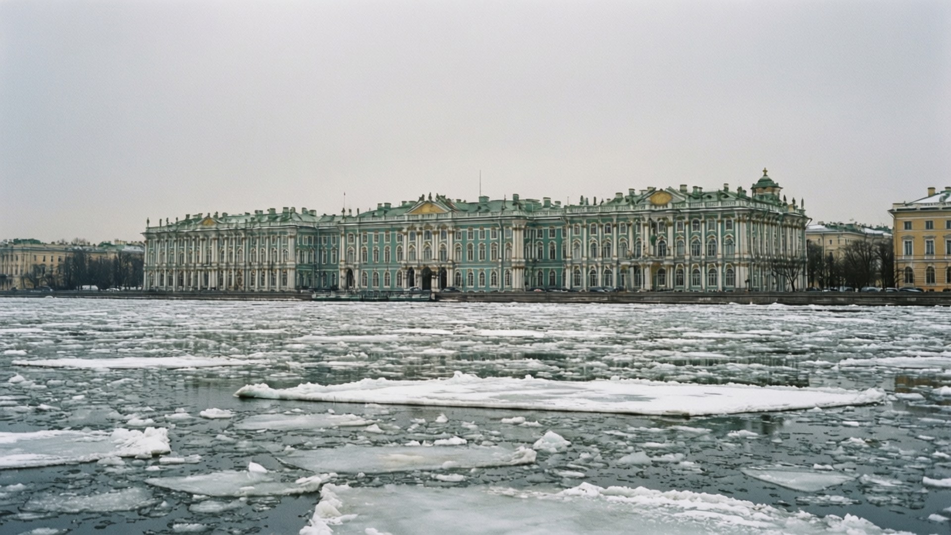 Winter Palace in Gray 壁紙 — 一座宏偉的新古典主義宮殿，擁有綠色和白色的外牆，矗立在結冰的河流後面，河面上滿是破碎的浮冰，天空多雲。