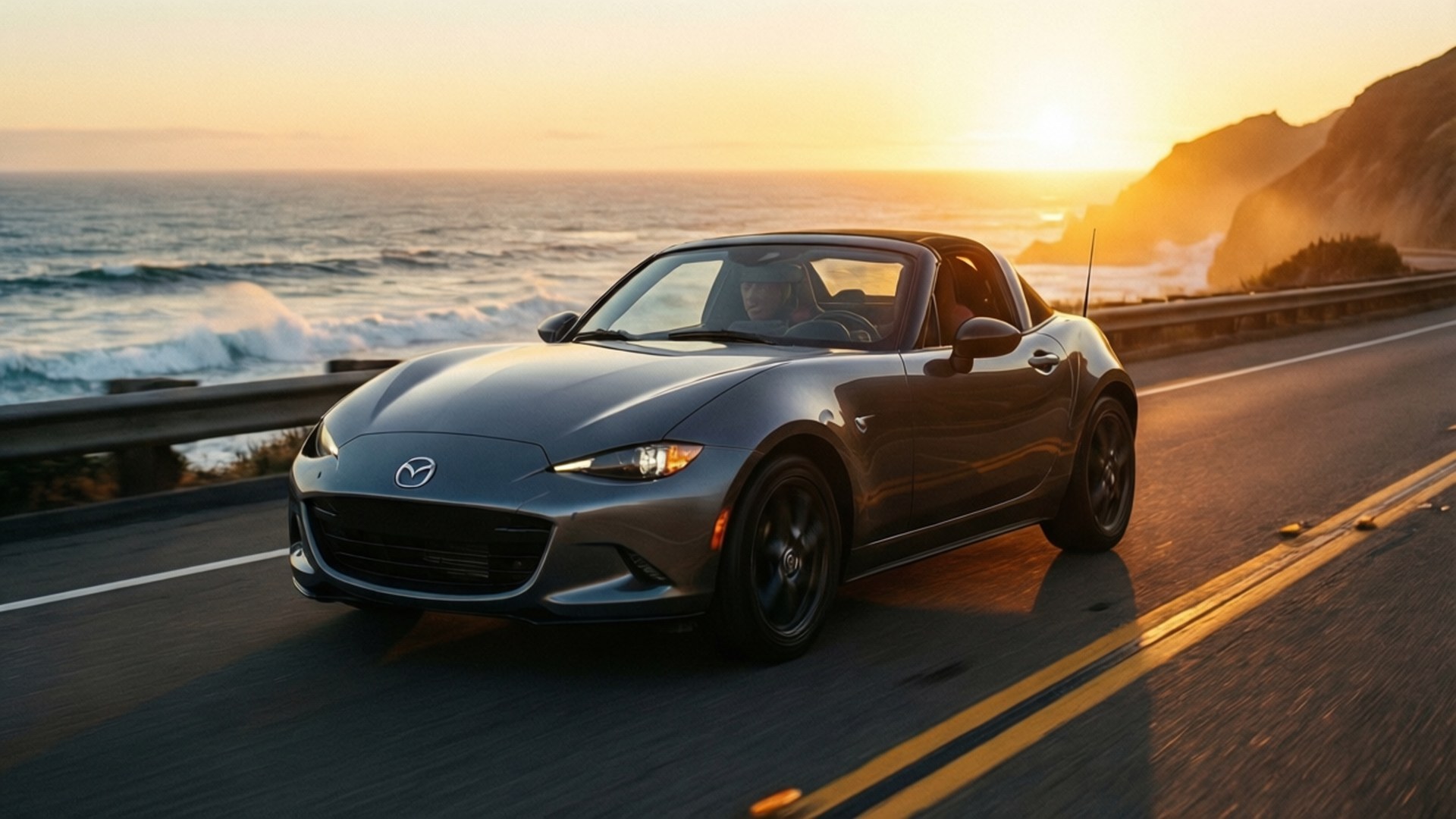 Coastal Sunset Miata 壁紙 — 一架時尚的灰色馬自達Miata敞篷車沿著海岸公路巡航，背景是金色夕陽、拍岸海浪和懸崖。