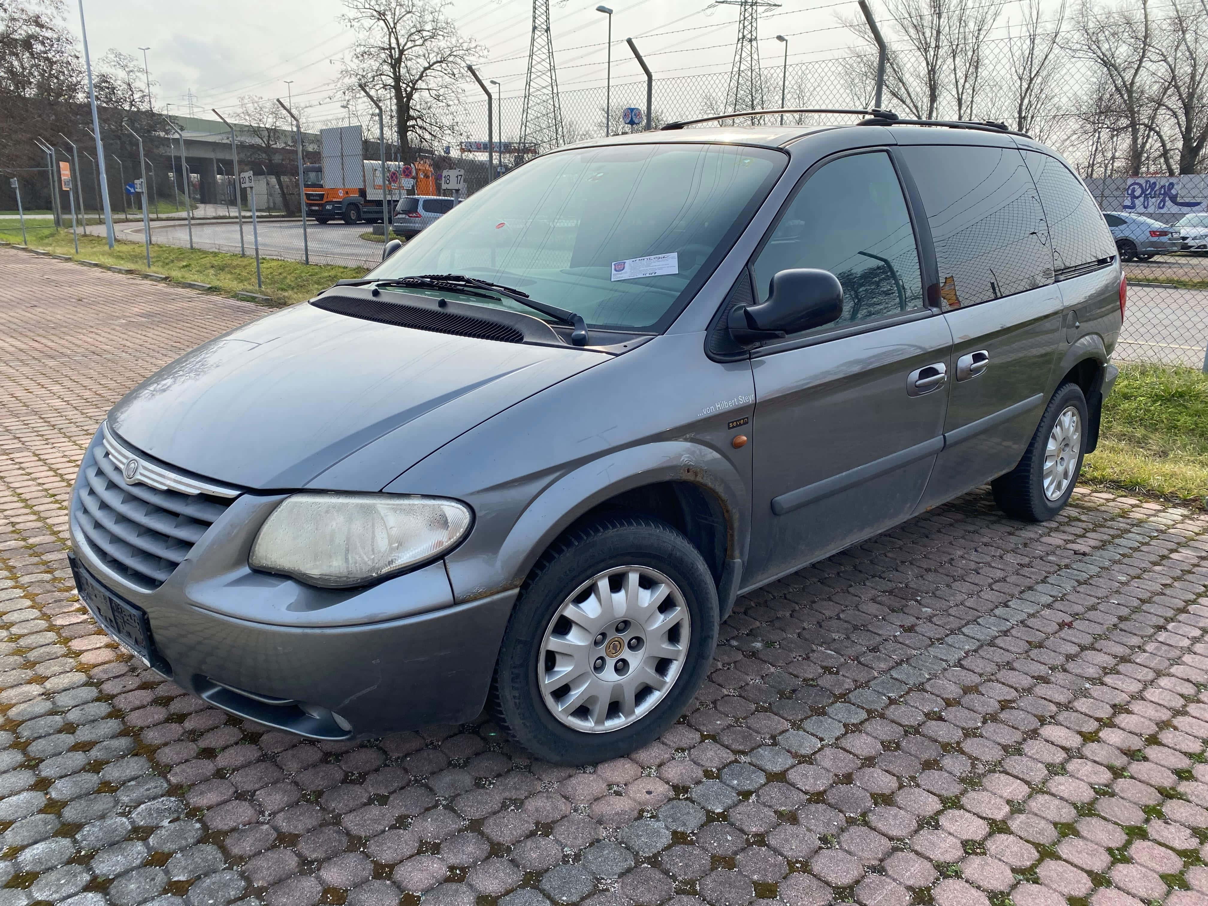 Chrysler Voyager 桌布 壁紙 — 一輛灰色小型貨車停放在濕漉漉的鵝卵石地上，天空多雲，背景有光禿禿的樹木和工業圍欄。