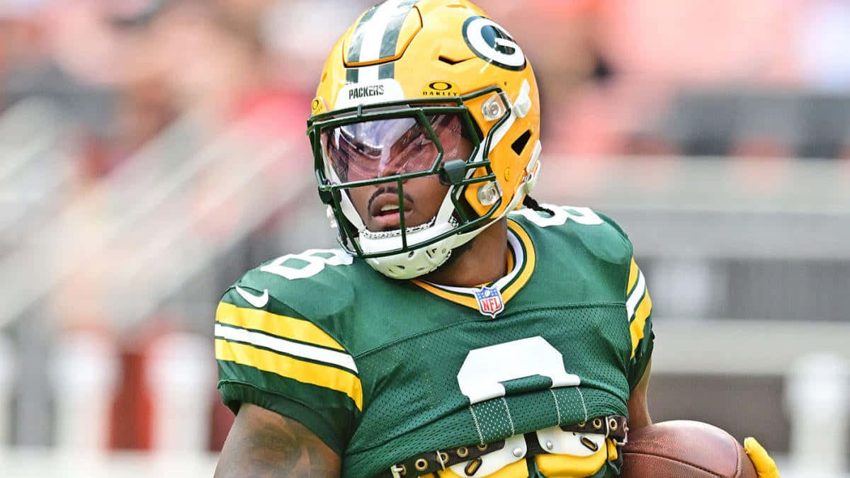  壁紙 — 一位綠灣包裝工美式足球員身穿綠色和金色制服，在日間比賽中手持足球，背景中的體育場看台模糊不清。