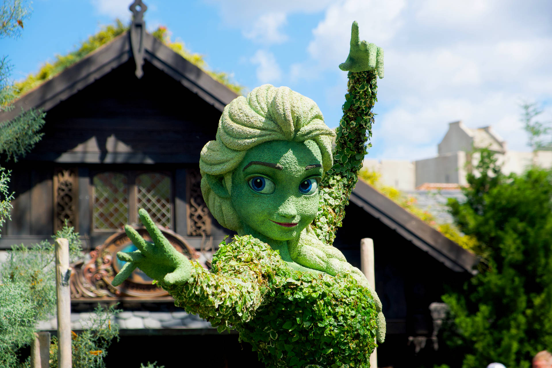 《冰雪奇緣》中艾莎的綠色修剪植物雕塑在木建築前揮手。 — 迪士尼公主 壁紙