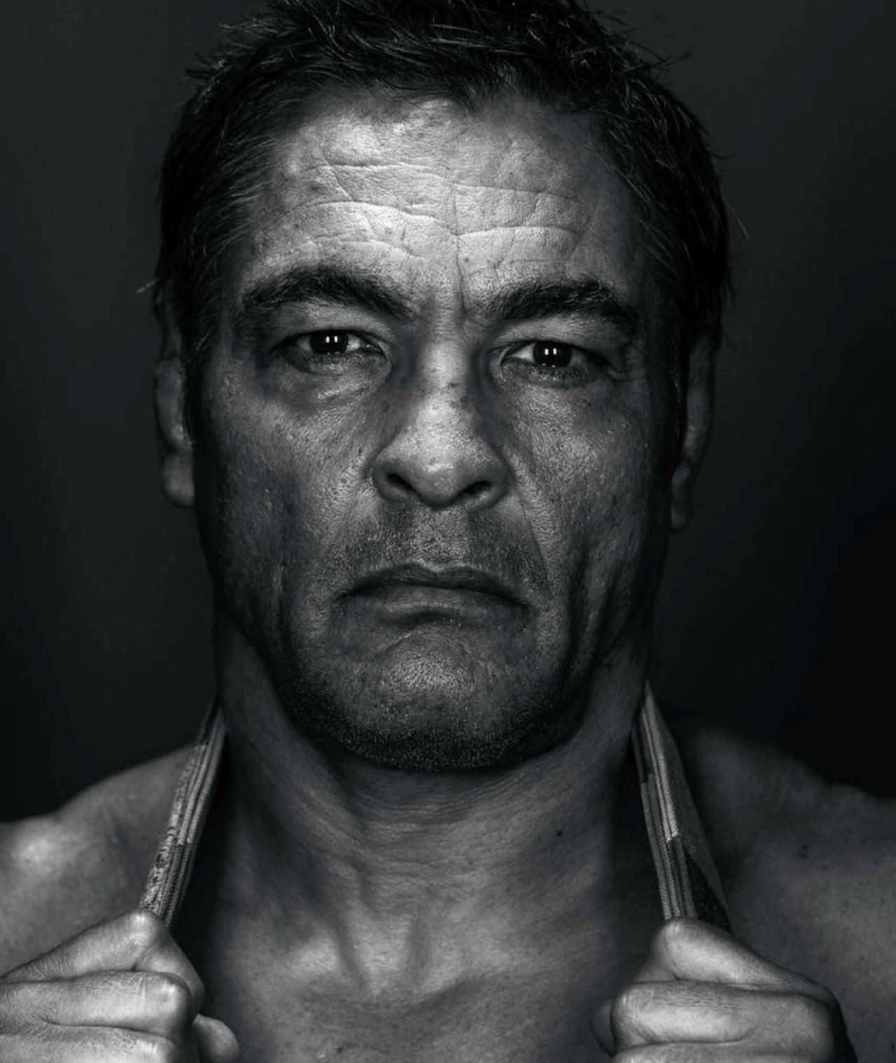 Rickson Gracie 桌布 壁紙 — 一個粗糙的黑白肖像，一個肌肉發達的男人帶著強烈的眼神，拉起他的背心露出胸膛。