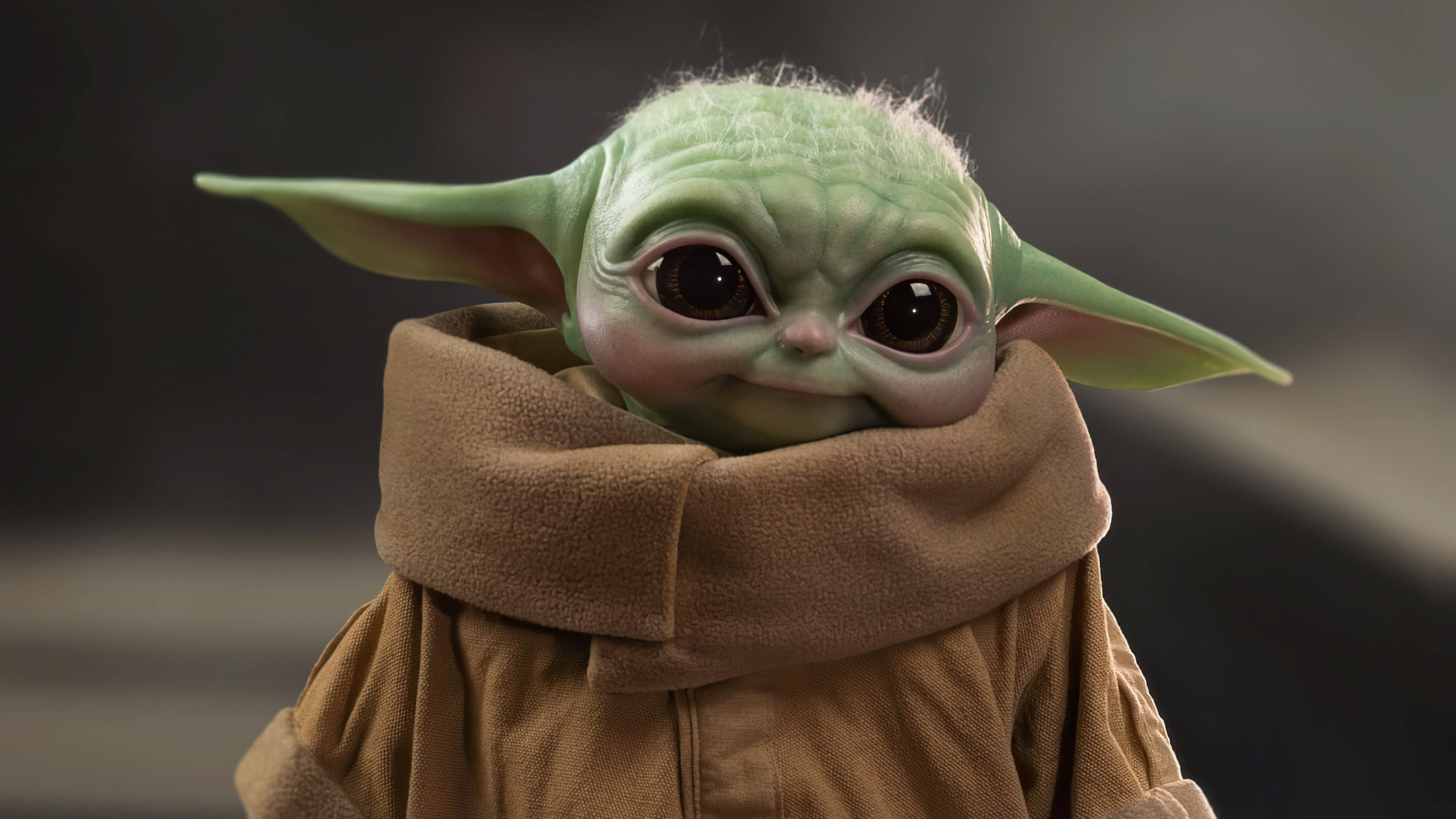 Mandalorian And Baby Yoda 桌布 壁紙 — Grogu 的特寫，他被棕色長袍包裹，有一雙大而富有表情的眼睛和尖耳朵，背景是模糊的灰色。