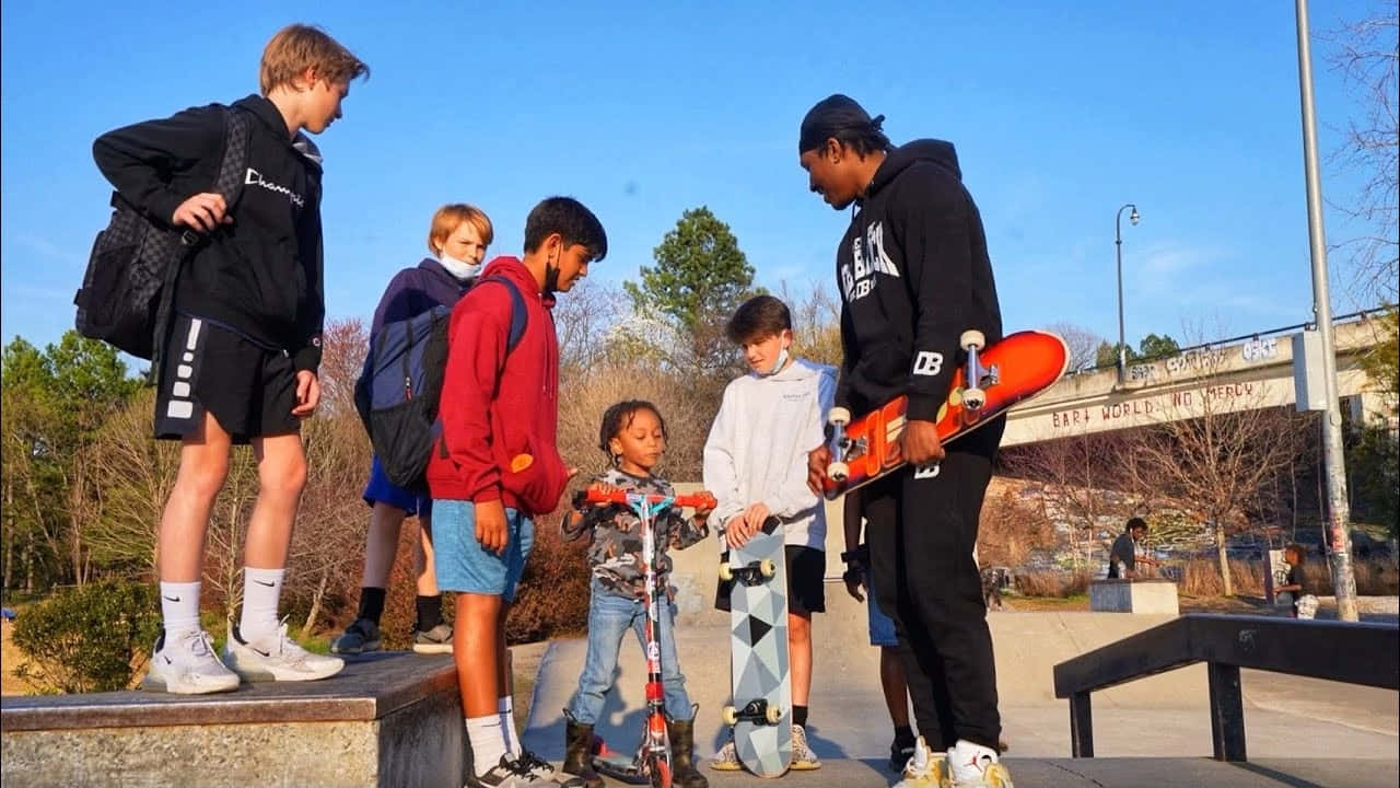 Duke Dennis 桌布 壁紙 — 一群多元化的青少年和一個小孩聚集在滑板和滑板車周圍，背景是陽光明媚的城市滑板公園，有光禿禿的樹木和一座橋。