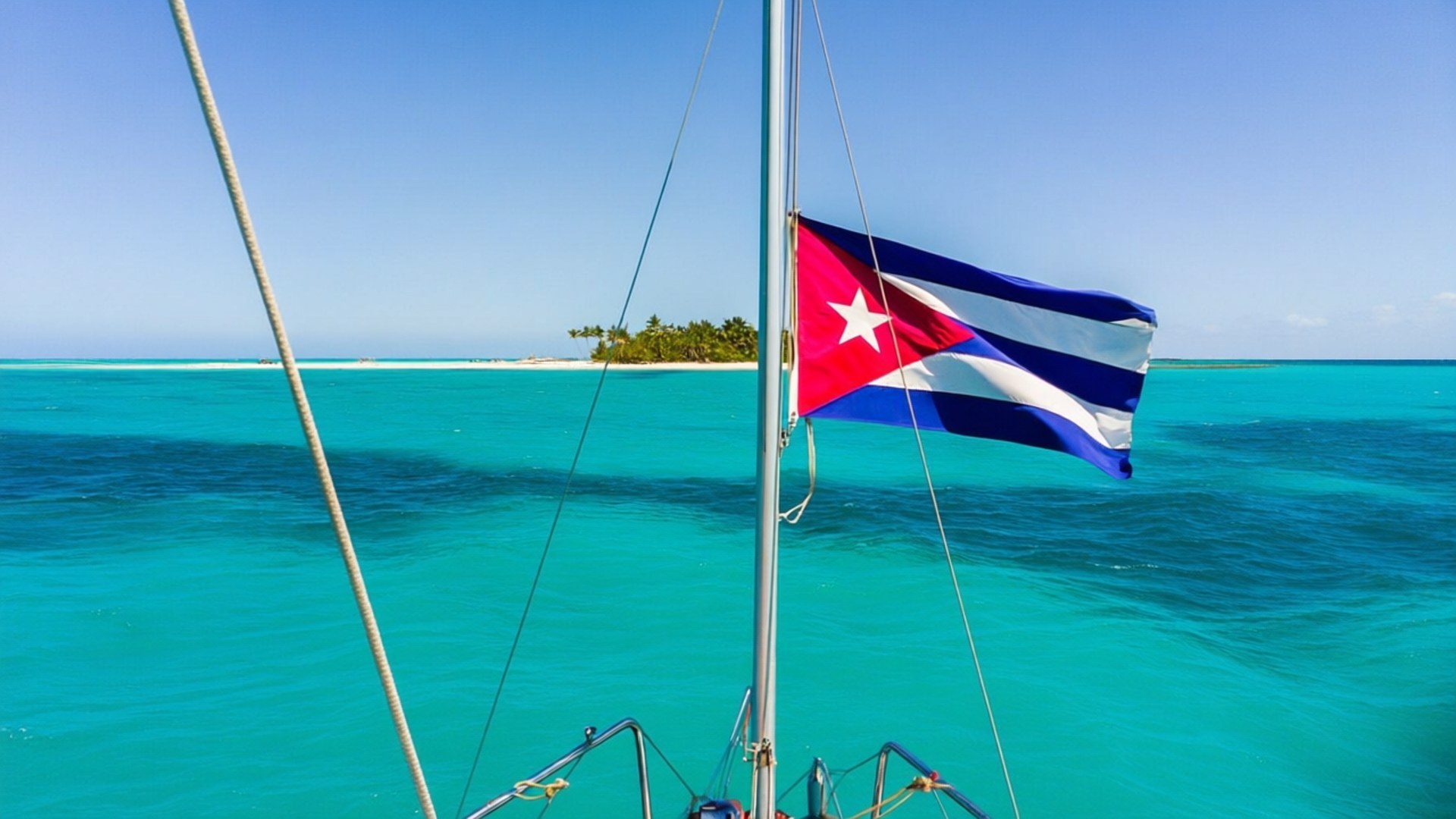Sailing Under Cuban Flag 壁紙 — 古巴國旗在帆船桅杆上飄揚，映襯著碧綠海水，遠處有熱帶島嶼，天空清澈湛藍。
