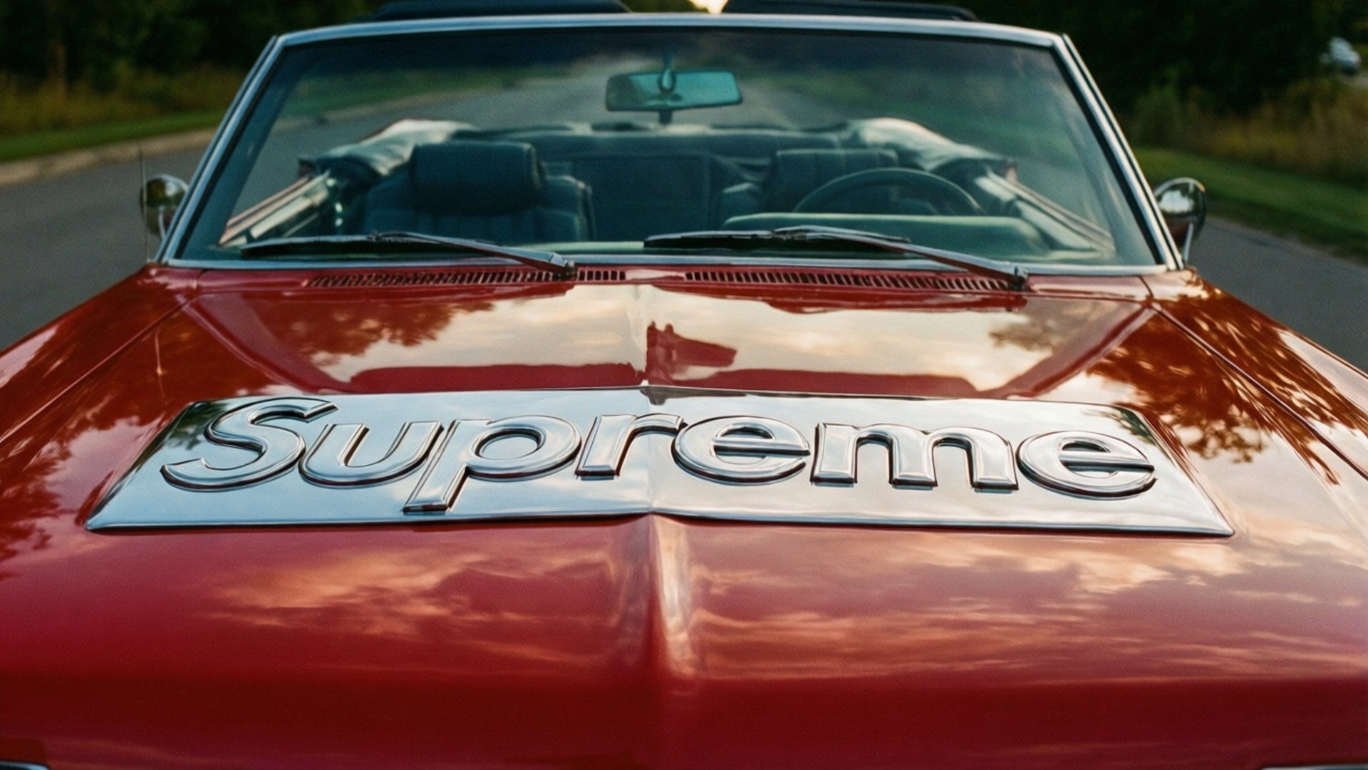 Supreme Vintage Car 壁紙 — 一輛光亮的紅色敞篷跑車，引擎蓋上有一個鍍鉻的Supreme標誌，反射著多雲的天空。