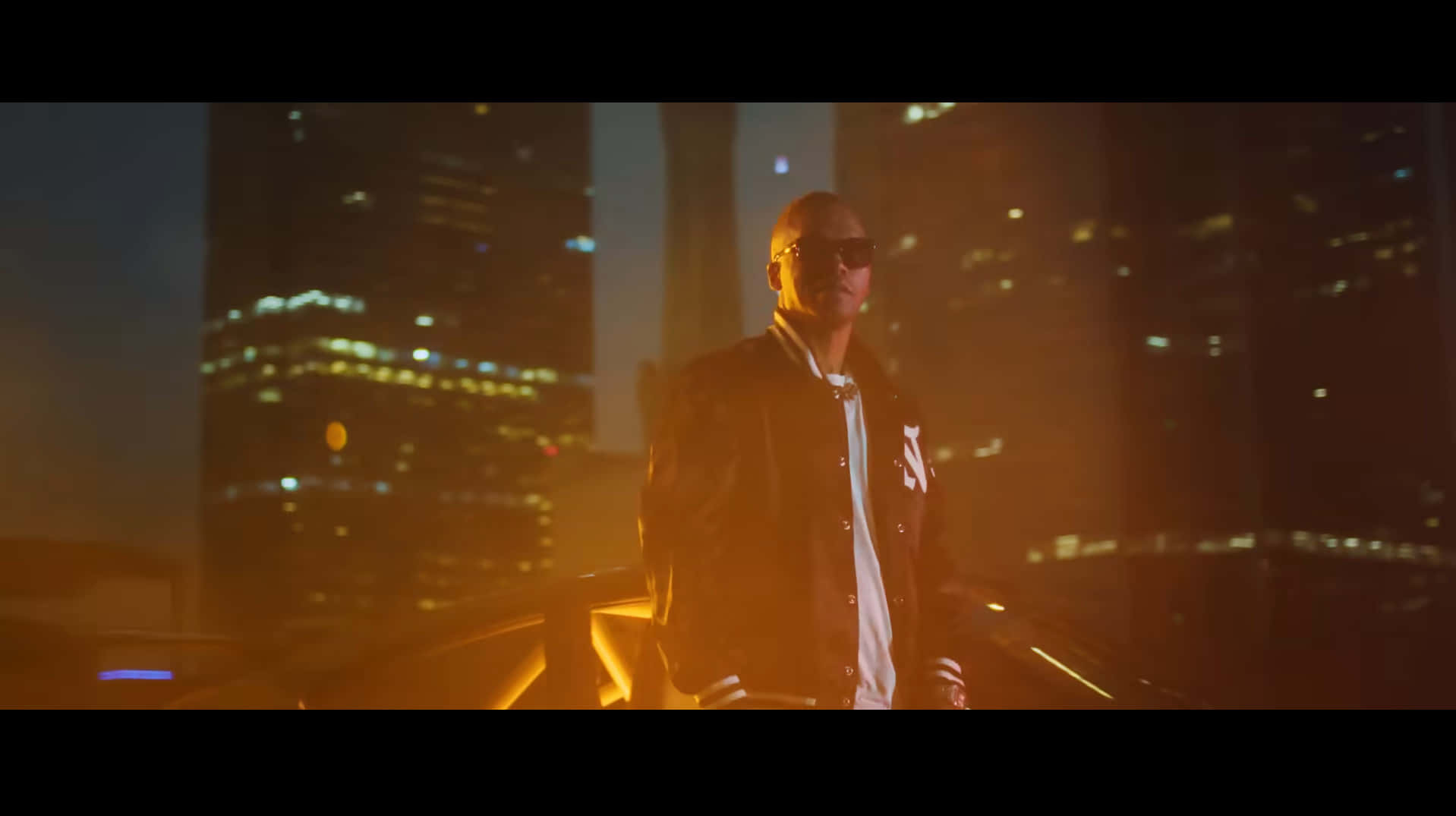  壁紙 — 一個光頭男子戴著太陽眼鏡和棒球外套，在夜晚的屋頂上自信地站著，背後是閃爍的城市天際線。