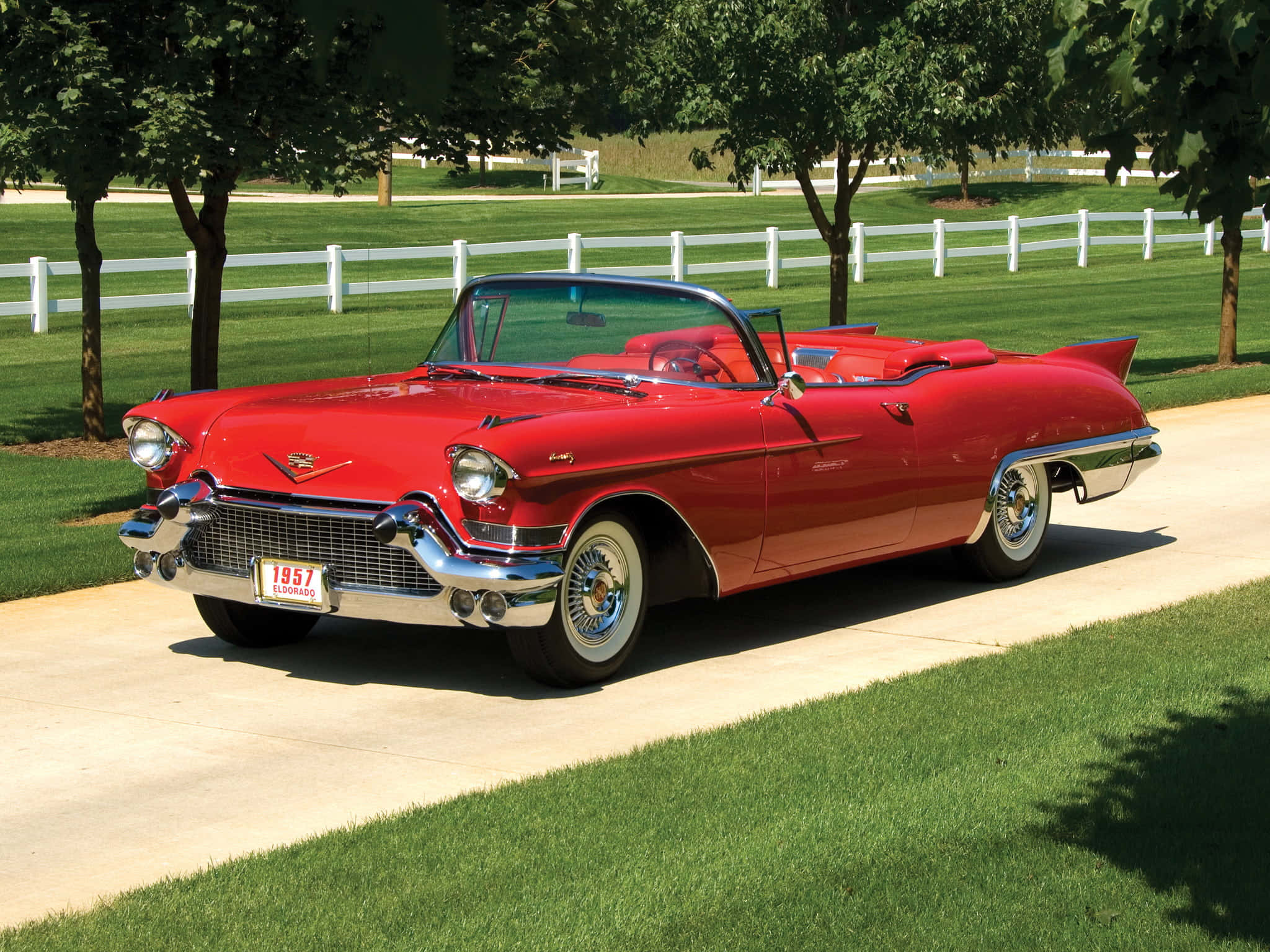 Cadillac Eldorado 桌布 壁紙 — 一輛光澤的紅色1957年凱迪拉克敞篷車停泊在鋪砌的小路上，旁邊是茂密的綠草和白色圍欄，在明亮的陽光下。